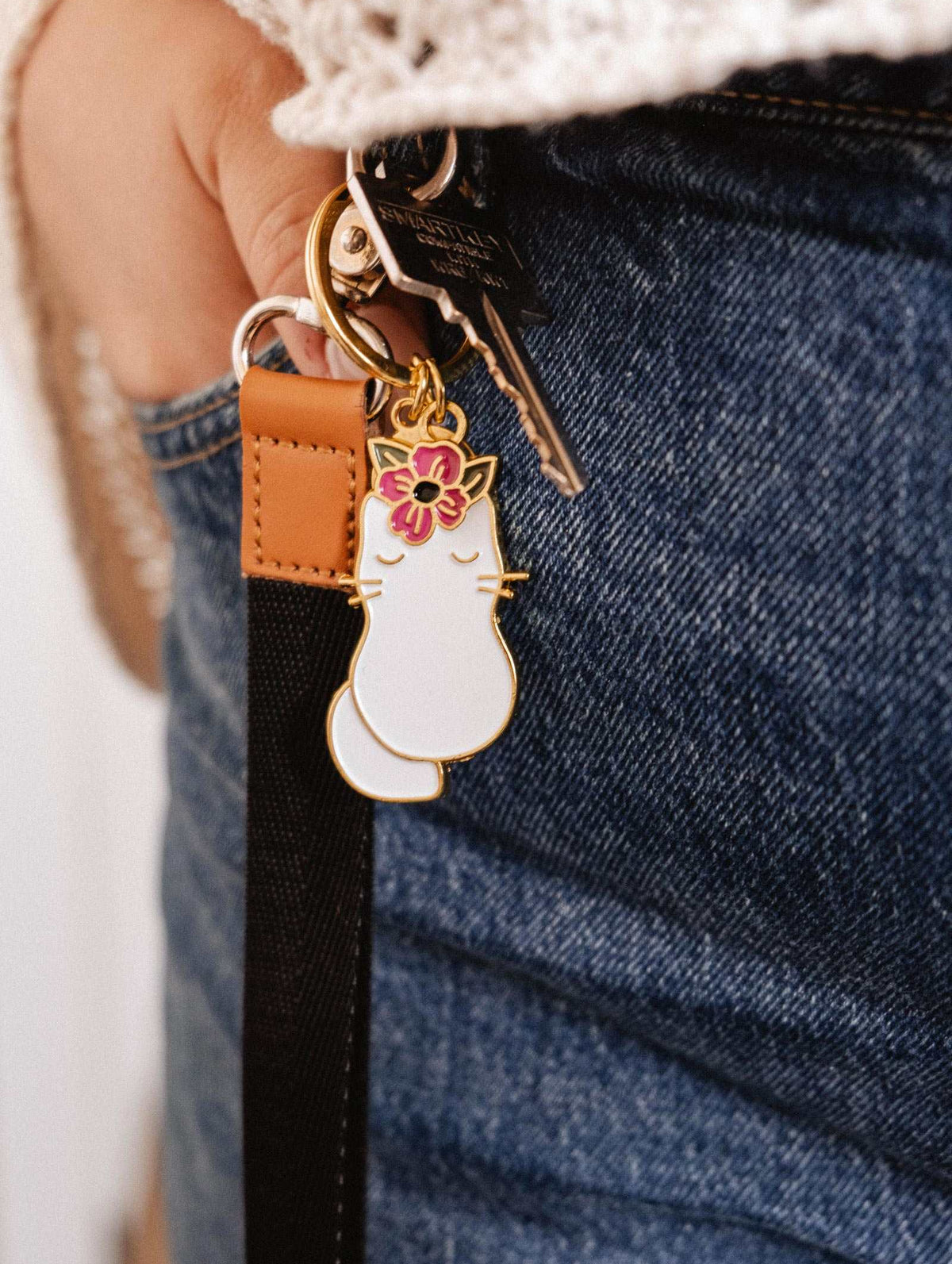 Gros plan d'une personne en jeans avec le Porte-clés Chat Fleuri de Mimi & August — un chat blanc avec une fleur rose — accroché à des clés sur une boucle de ceinture, en faisant l'accessoire parfait pour les amoureux des chats.