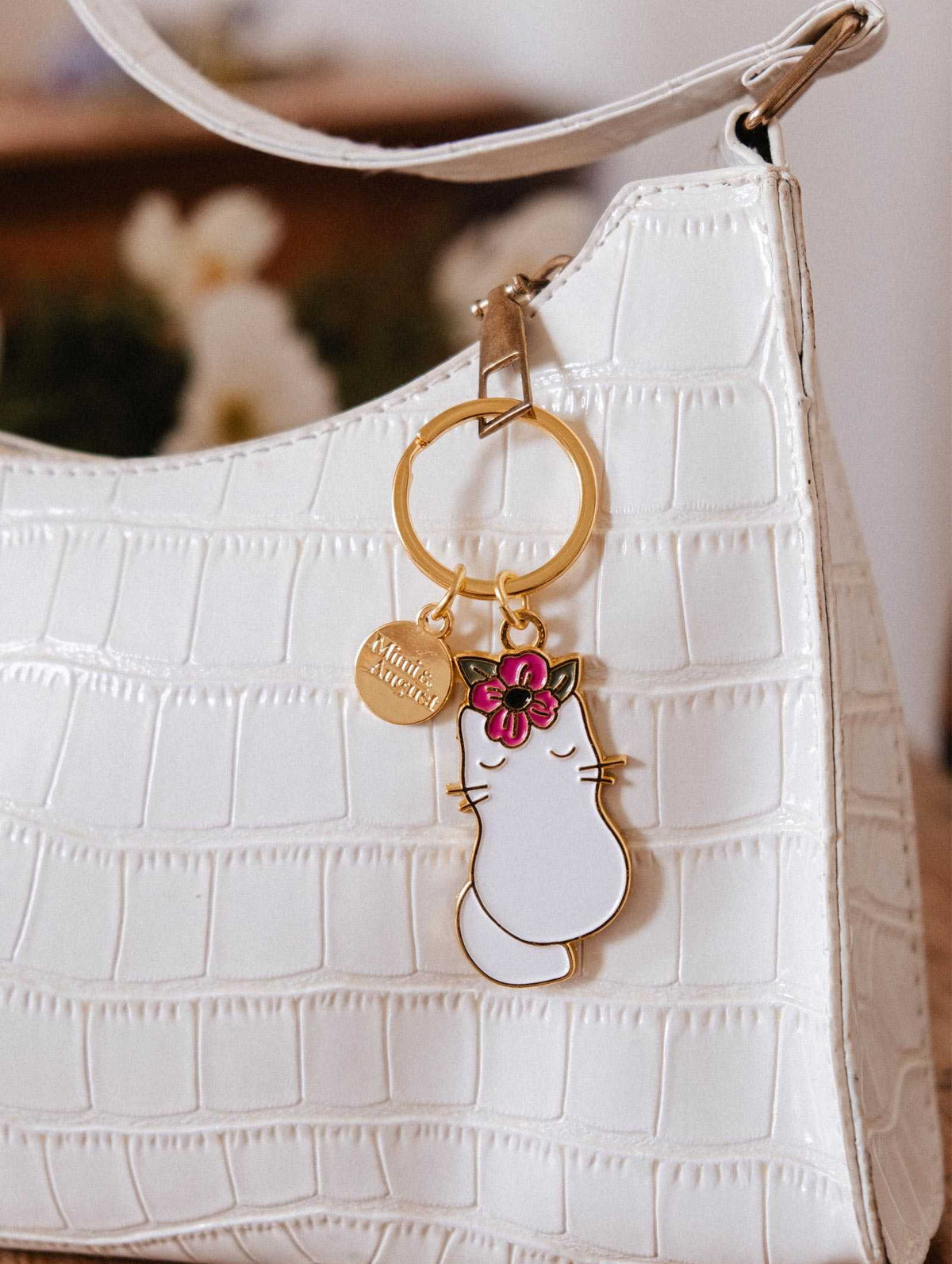 Le Porte-clés Chat Fleuri de Mimi & August, avec un charme de chat orné d'une fleur rose et un médaillon rond, est l'accessoire parfait pour les amoureux des chats à associer à votre sac à main blanc texturé.