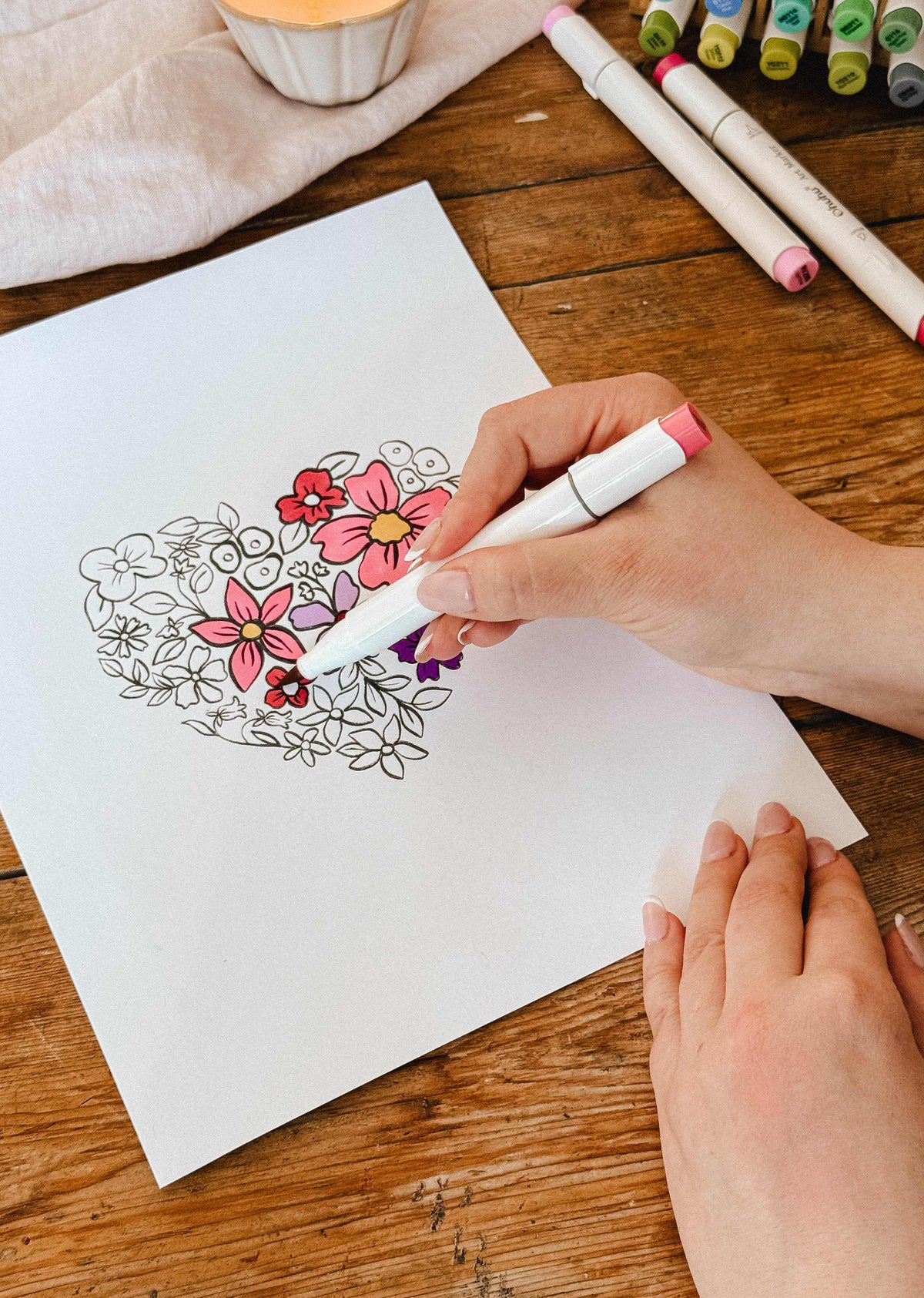 Someone colors the Mimi & August Floral Heart Coloring Sheet on white paper with pink and purple markers at a wooden table, enjoying a relaxing moment with this free coloring sheet.