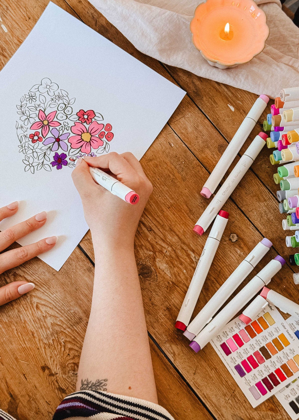 A person colors the Mimi & August Floral Heart Coloring Sheet with markers, surrounded by colorful markers, a color chart, and a lit candle—a perfect printable or free digital download for your next relaxing art session.