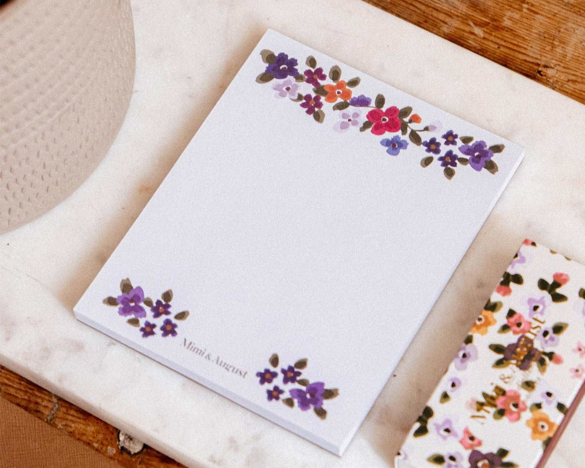 Mini Bloc-notes floral Bouquet de Mimi & August avec un stylo assorti sur une surface en marbre à côté d'une plante et d'une table en bois.