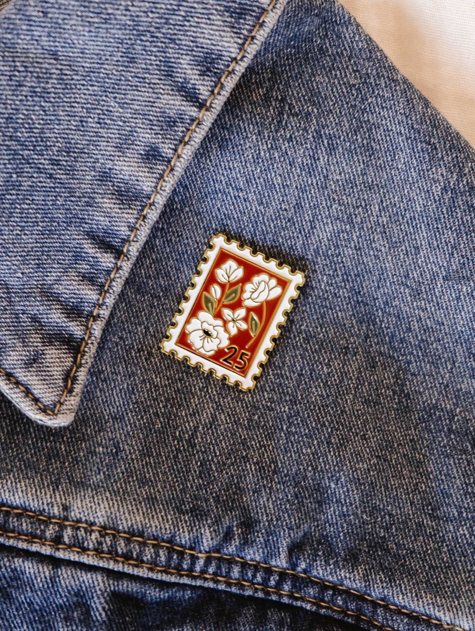 The Mimi & August Floral Stamp Enamel Pin, shaped like a vintage stamp with a floral charm and the number 25, is attached to a blue denim jacket.