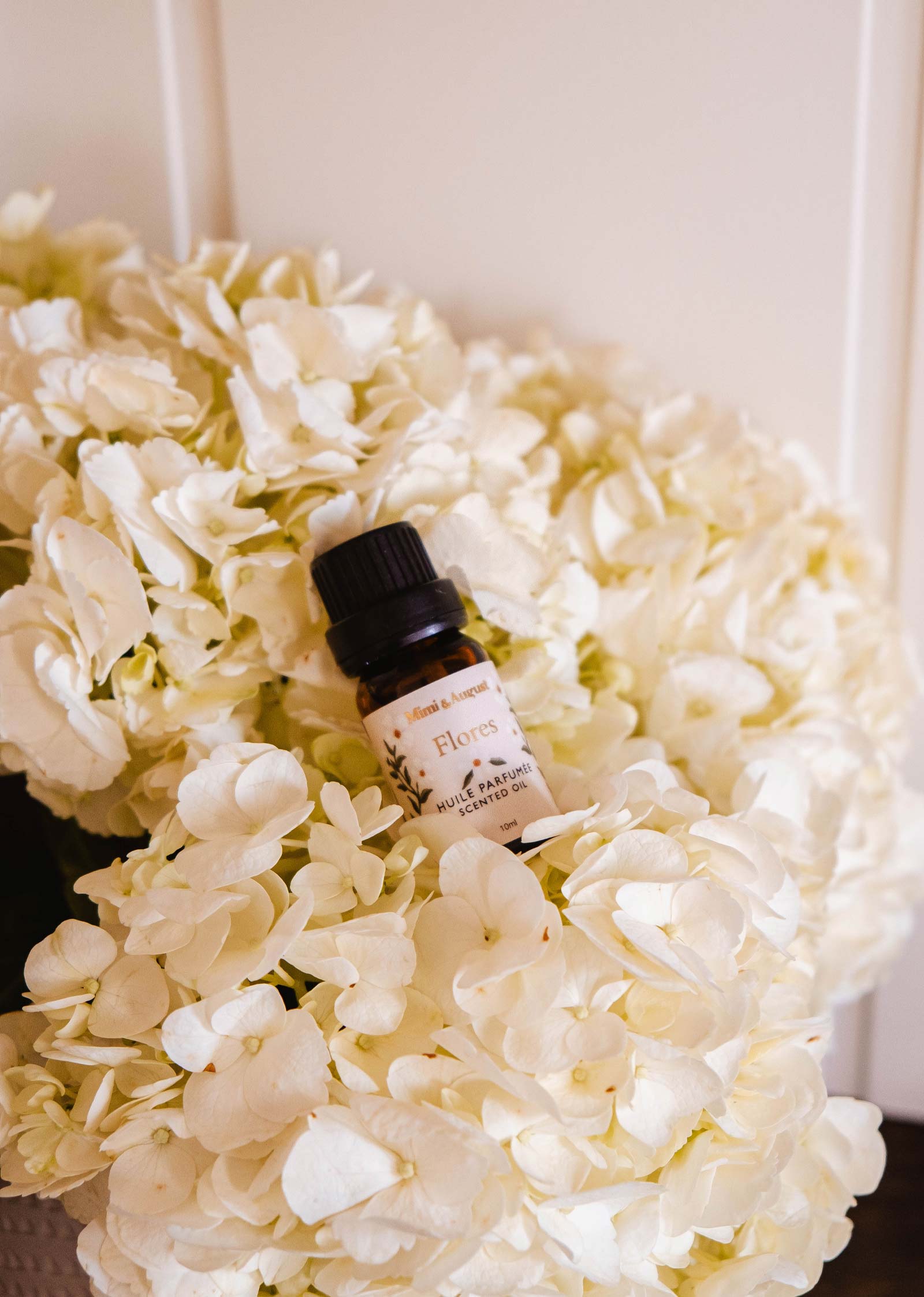 A small bottle of Mimi & August Scented Oil - Flores sits atop white hydrangeas, ideal for bringing a touch of home fragrance to any space against a light background.