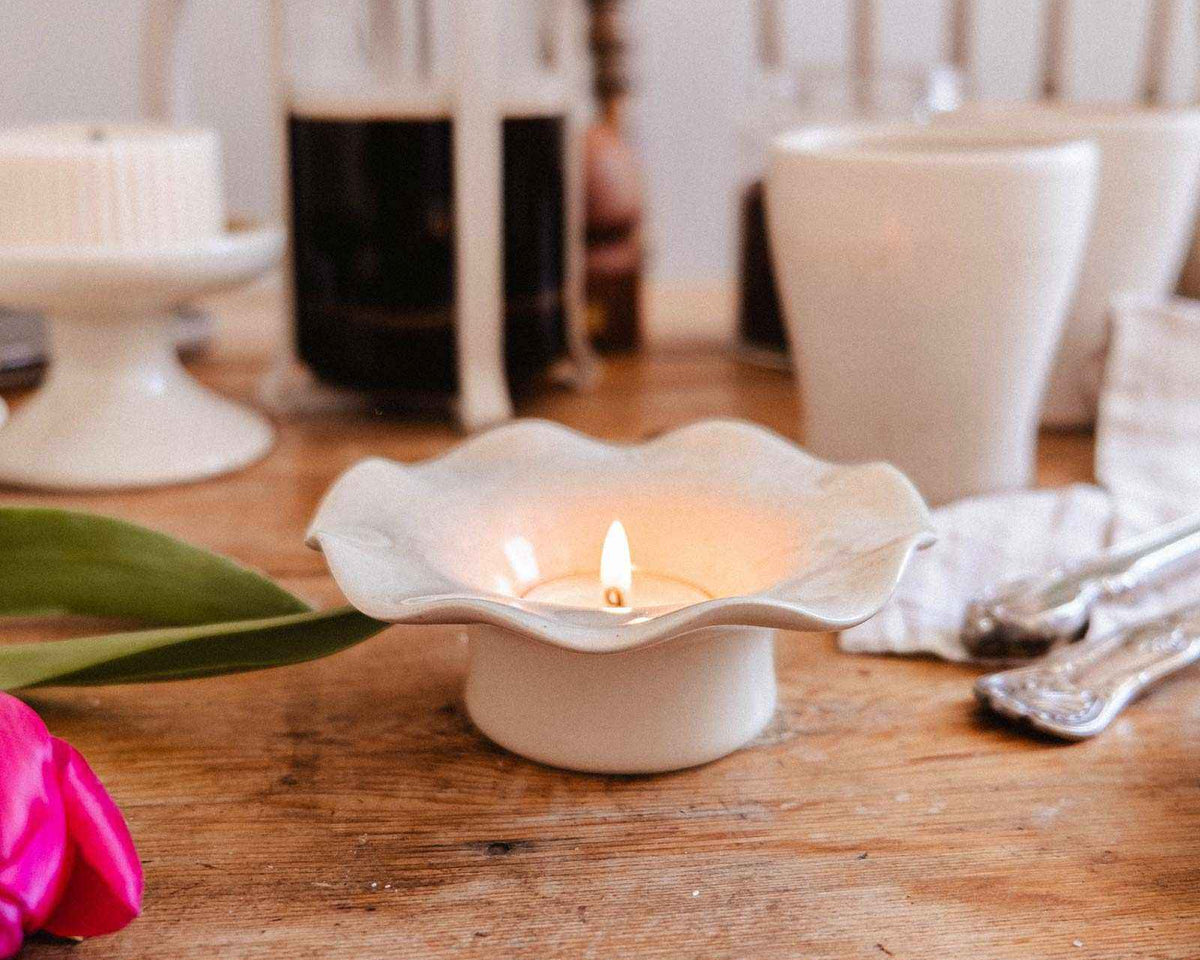 Une bougie allumée dans le Bougeoir en céramique cannelé de Mimi & August repose sur une table en bois avec une cafetière à piston, des tasses, des ustensiles et des tulipes roses.
