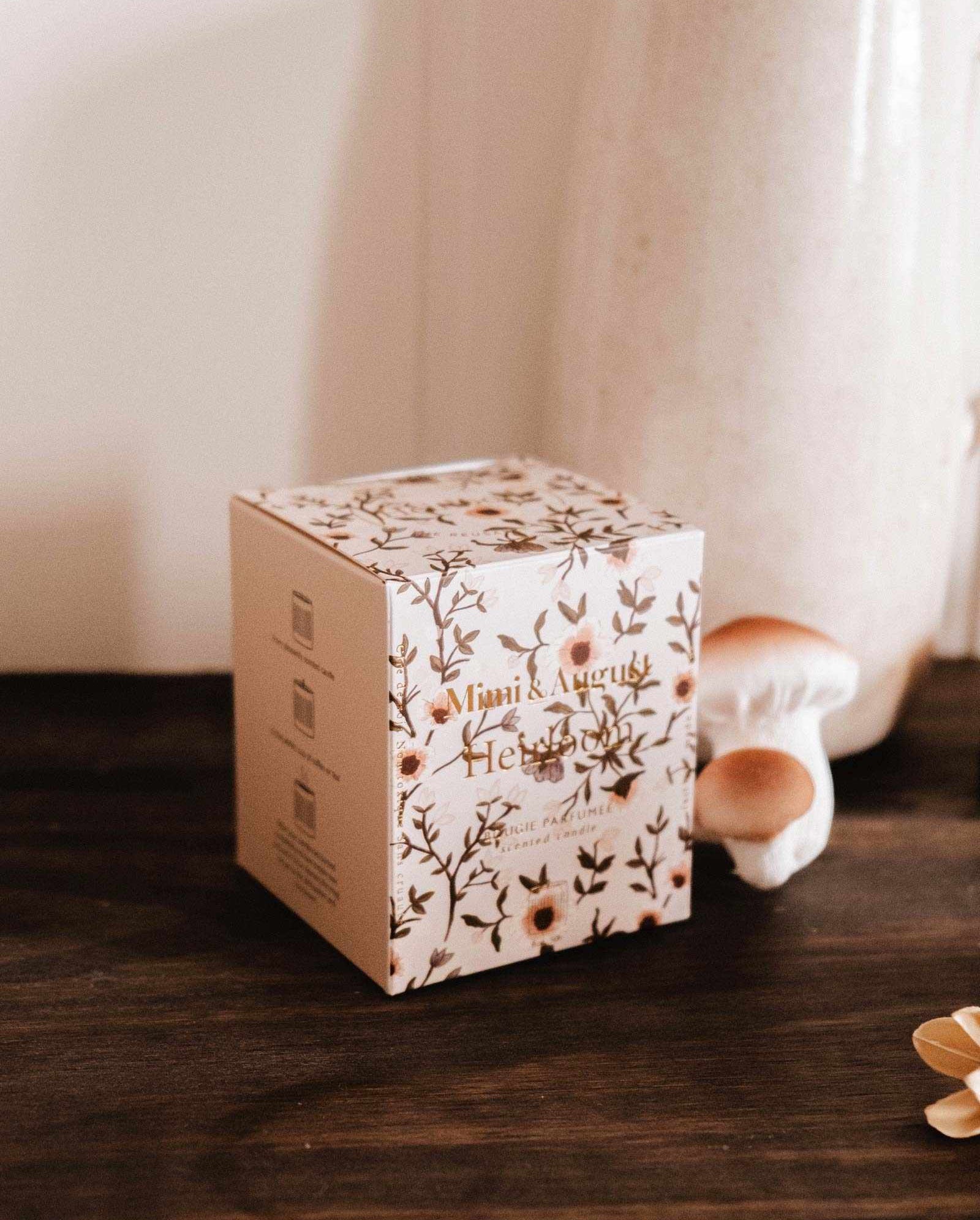 The Mimi & August Heirloom - Reusable Candle, in a decorative box with floral patterns and gold text, sits on a dark wooden surface beside a ceramic vase and two small mushroom ornaments.