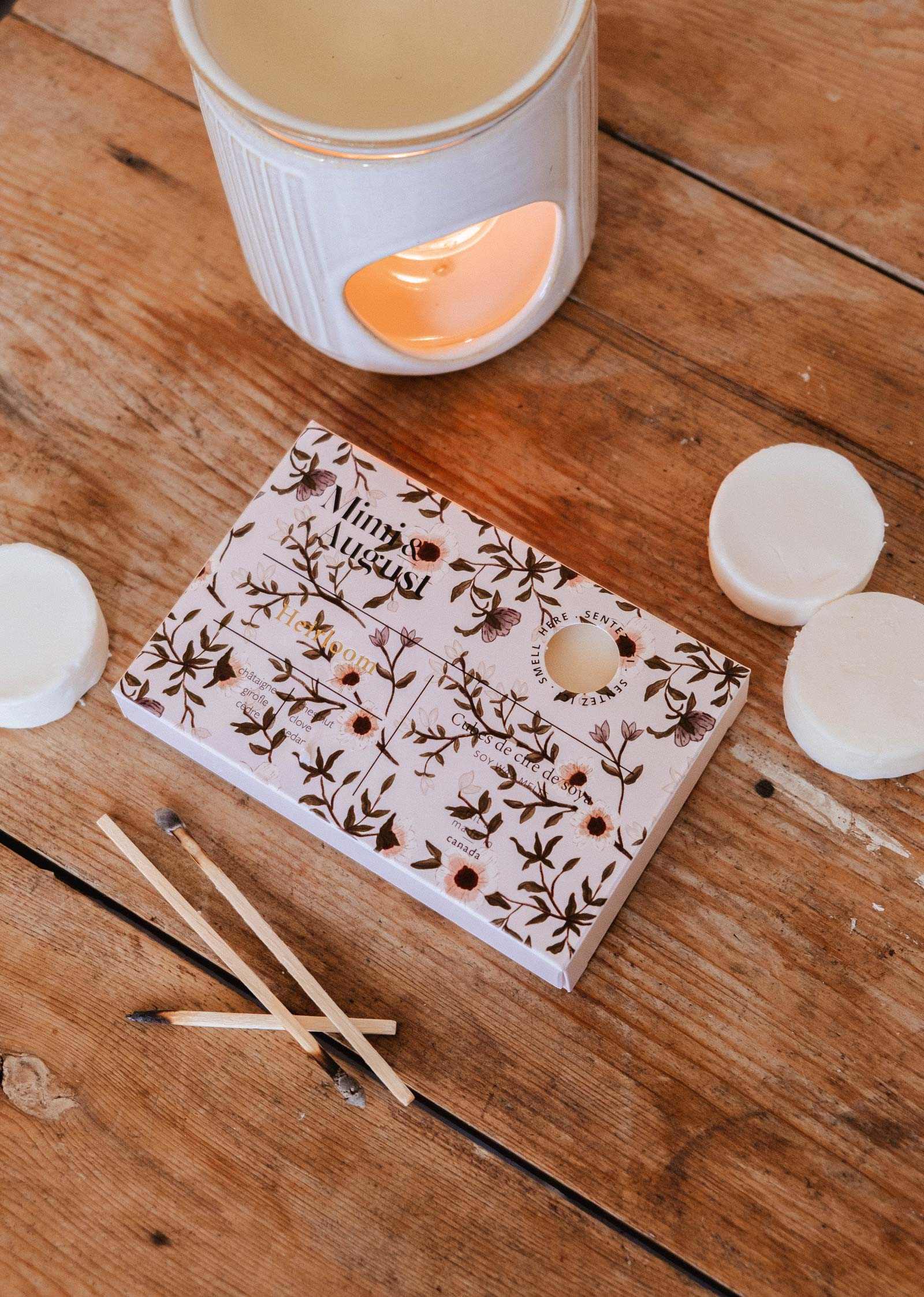 A Mimi & August Soy Wax Melts - Heirloom sits with a ceramic wax warmer on a wooden table, surrounded by floral packaging, three soy wax melts, and burnt matches—creating an inviting setting for natural fragrance.