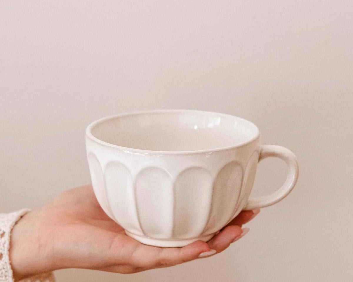 A hand holds a Ceramic Latte Mug by Mimi & August, displaying its white ceramic body with a textured, scalloped pattern against a plain light background.