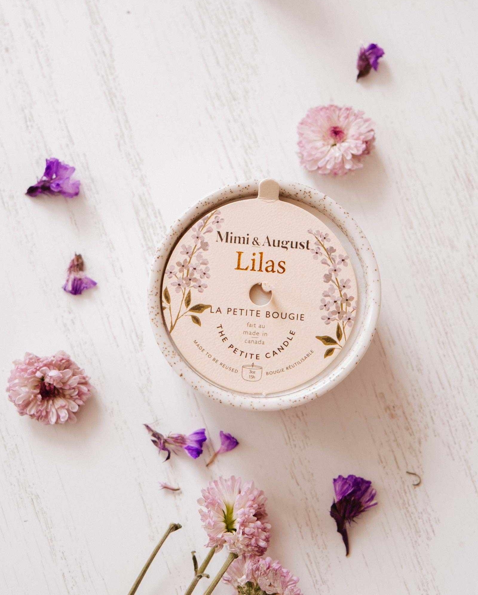 A Mimi & August Lilac - Reusable Candle sits on a white wooden surface, ideal for aromatherapy, surrounded by scattered pink and purple flowers.