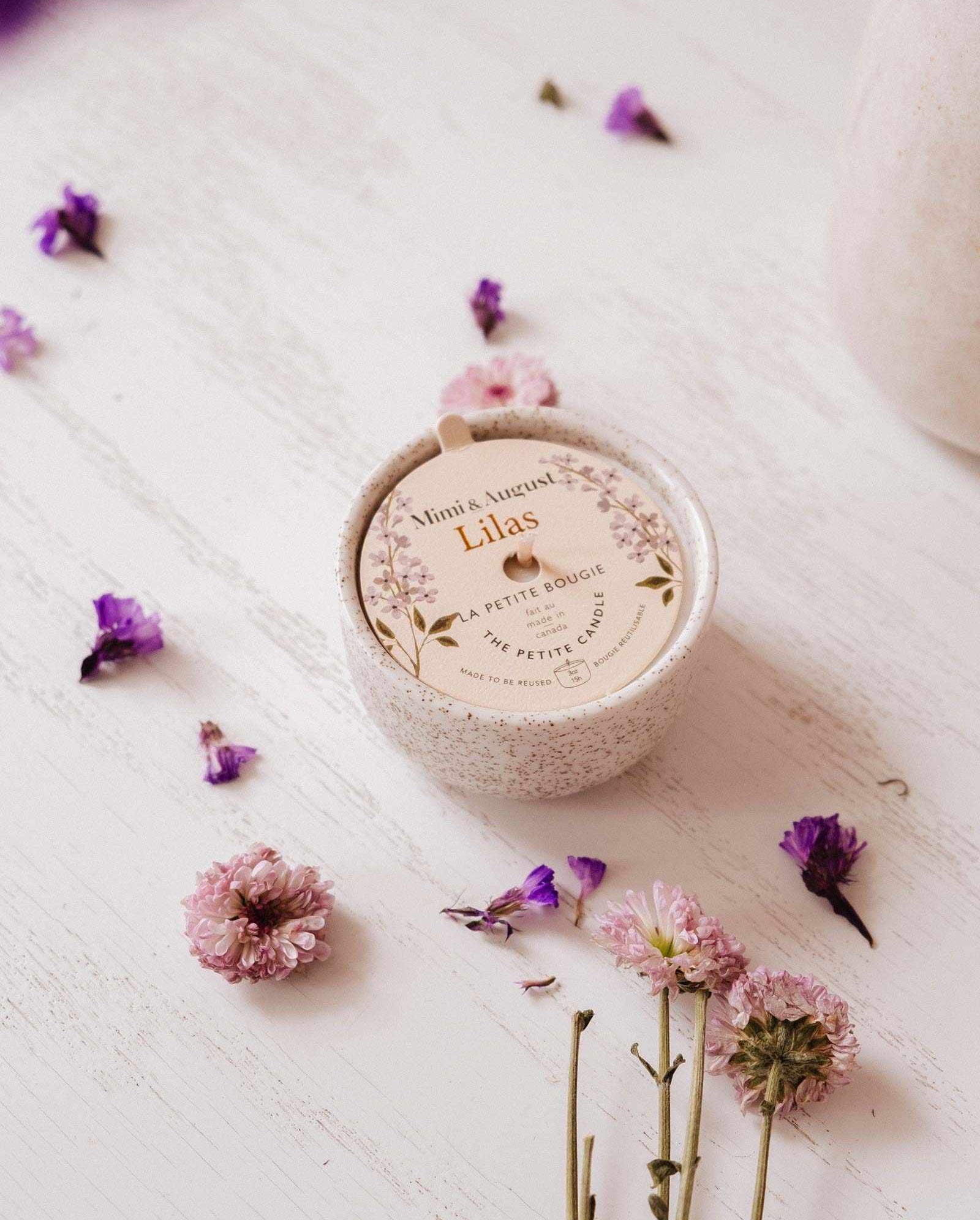 The Lilac - Reusable Candle by Mimi & August rests on a white wooden surface, surrounded by pink and purple flowers, bringing elegant home decor and soothing lilac fragrance to the space.