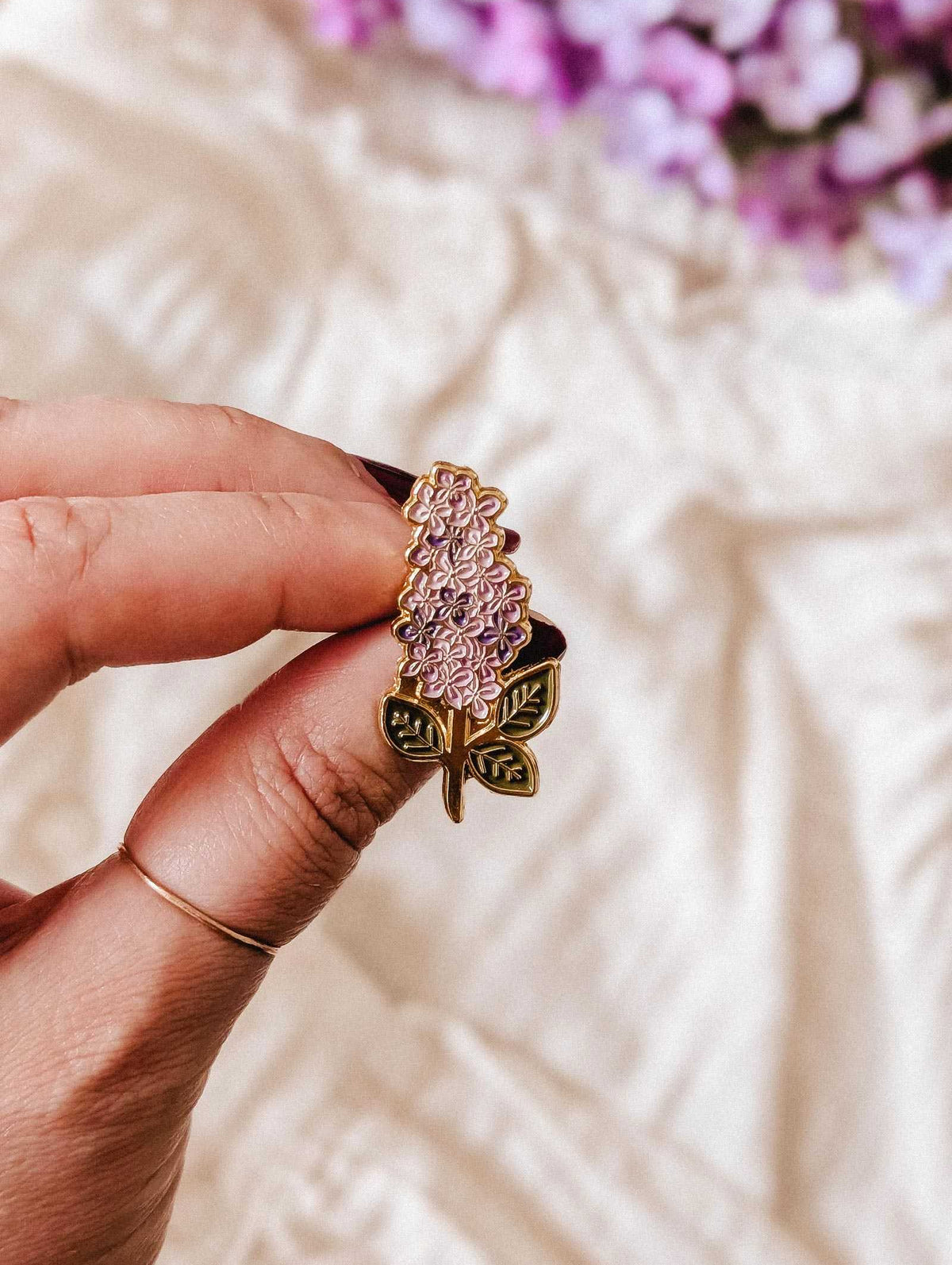 A hand holds the Mimi & August Lilac Enamel Pin, featuring gold tone brass with green leaves; a soft fabric and purple flowers are blurred in the background.