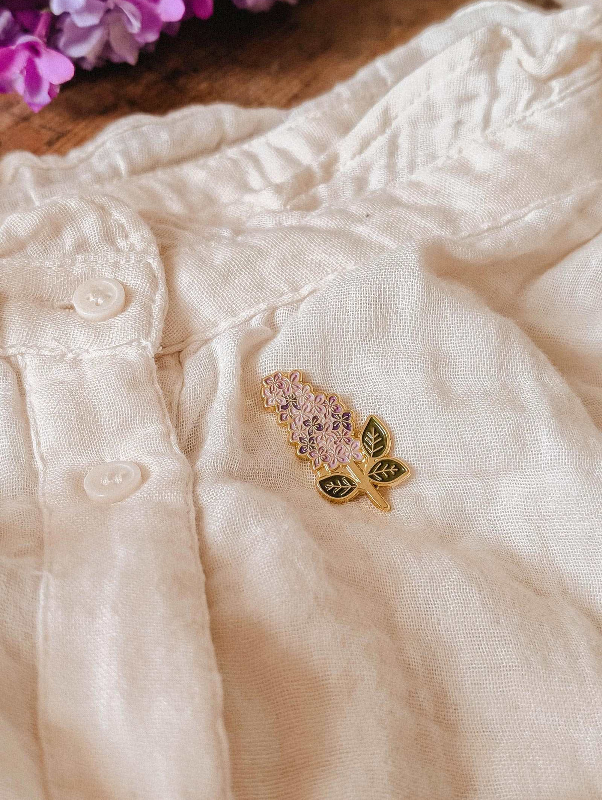The Mimi & August Lilac Enamel Pin in gold tone brass is attached to a cream linen garment with buttons, with lilac flowers visible in the upper left corner.
