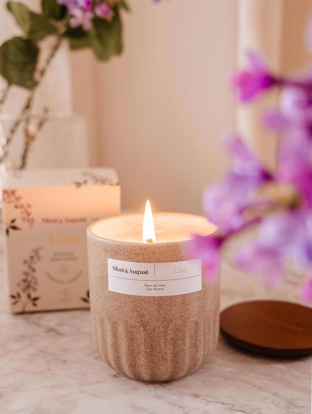 The Lilac - Reusable Candle by Mimi & August in a beige ceramic jar sits on marble, bringing aromatherapy and elegance to your home, with its box and purple flowers as a stylish backdrop.