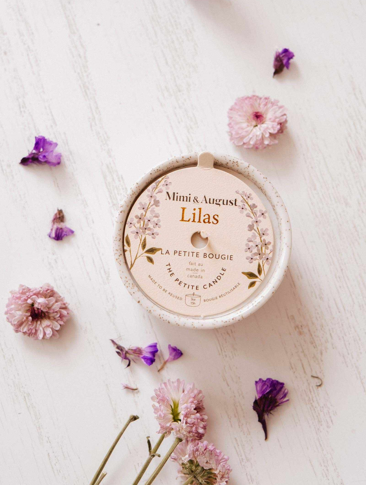 A Mimi & August Lilac - Reusable Candle sits on a white wooden surface, ideal for aromatherapy, surrounded by scattered pink and purple flowers.