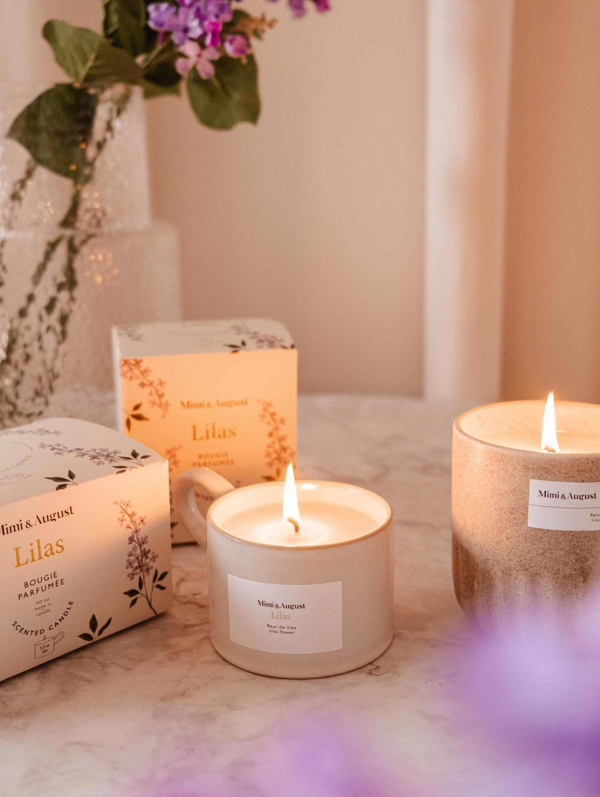 Two lit Mimi & August Lilac - Reusable Candles labeled "Lilas" sit on marble, with a vase of purple flowers in the background—ideal for adding soothing aroma and elegant home decor to your space.