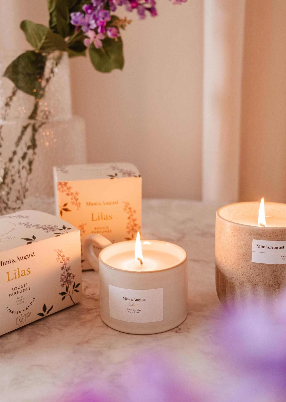 Two lit Mimi & August Lilac - Reusable Candles labeled "Lilas" sit on marble, with a vase of purple flowers in the background—ideal for adding soothing aroma and elegant home decor to your space.