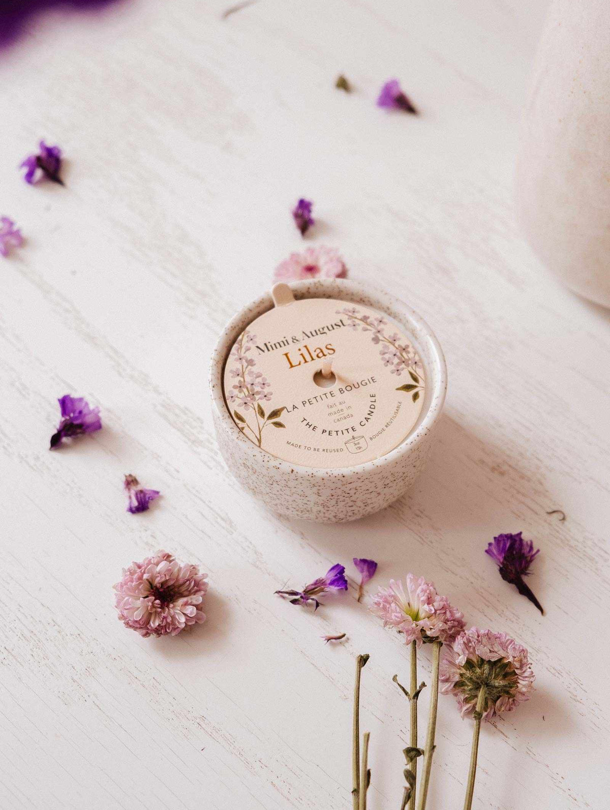 The Lilac - Reusable Candle by Mimi & August rests on a white wooden surface, surrounded by pink and purple flowers, bringing elegant home decor and soothing lilac fragrance to the space.