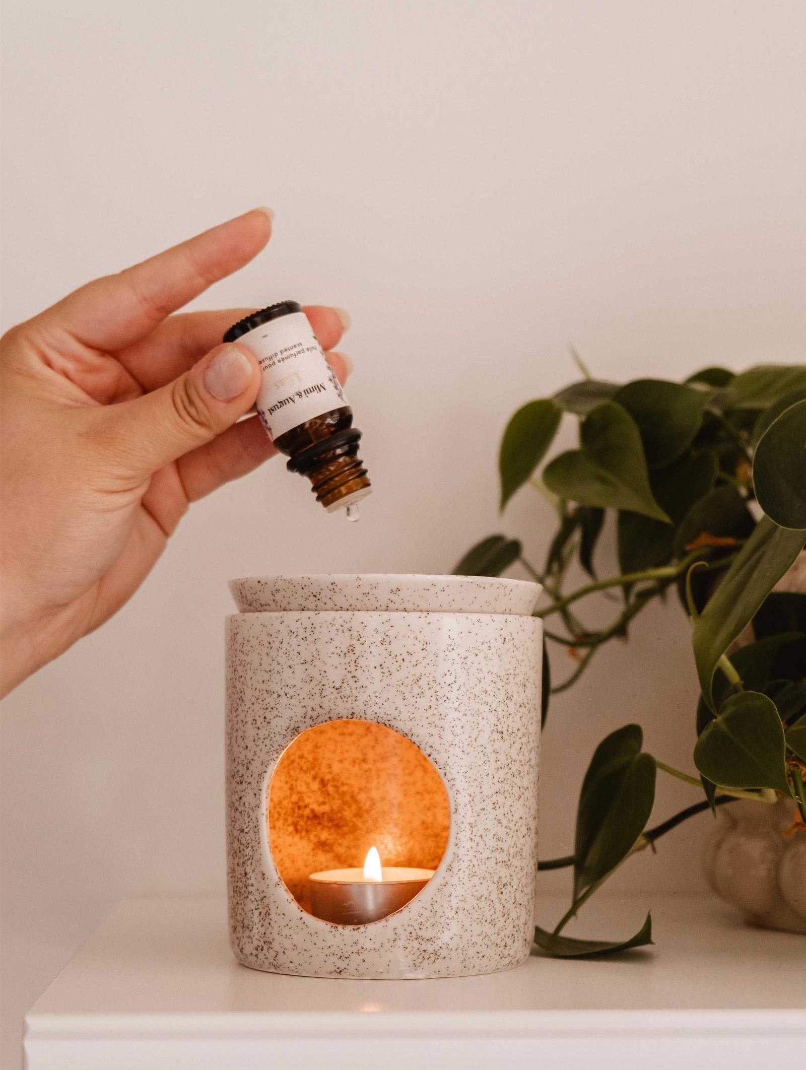 A hand is holding a small bottle of Scented Oil - Lilac by Mimi & August, adding a few drops into an aroma diffuser with a lit tealight candle inside. A green plant is visible in the background, enhancing the serene atmosphere with lilacs' soothing scent.