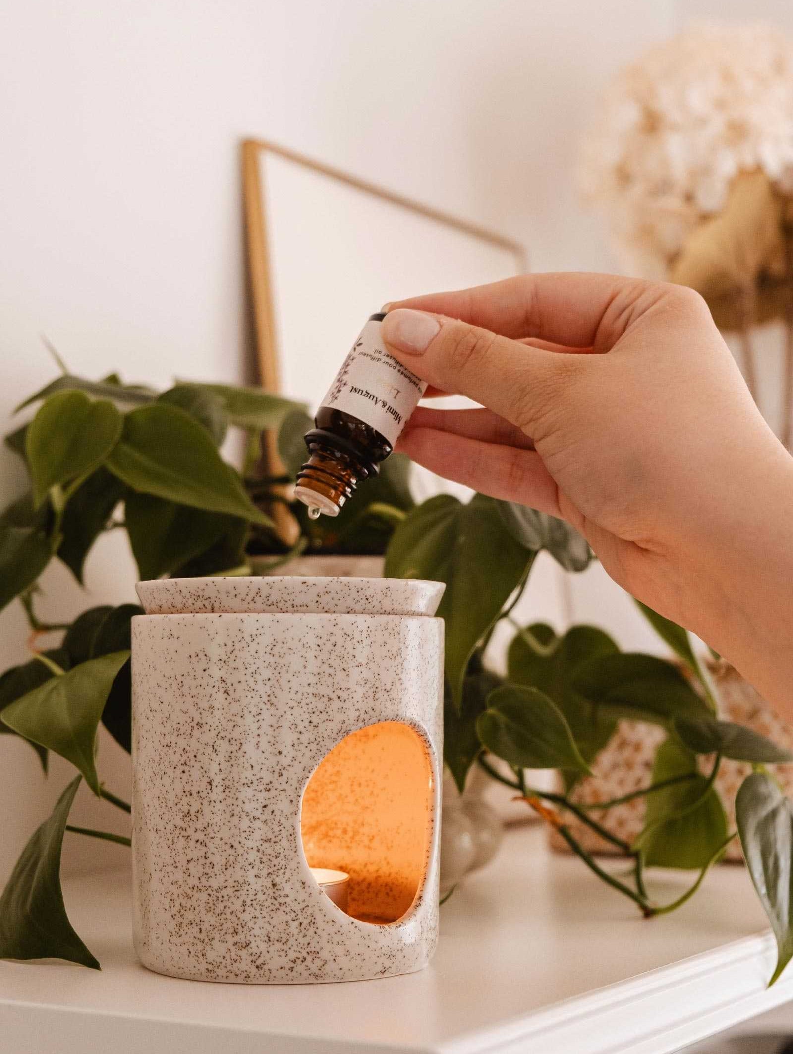 A hand is shown adding a few drops of Mimi & August Scented Oil - Lilac into a lit ceramic diffuser, set against a backdrop of green leaves and blurred decor items, creating an ambiance reminiscent of lilacs in spring.