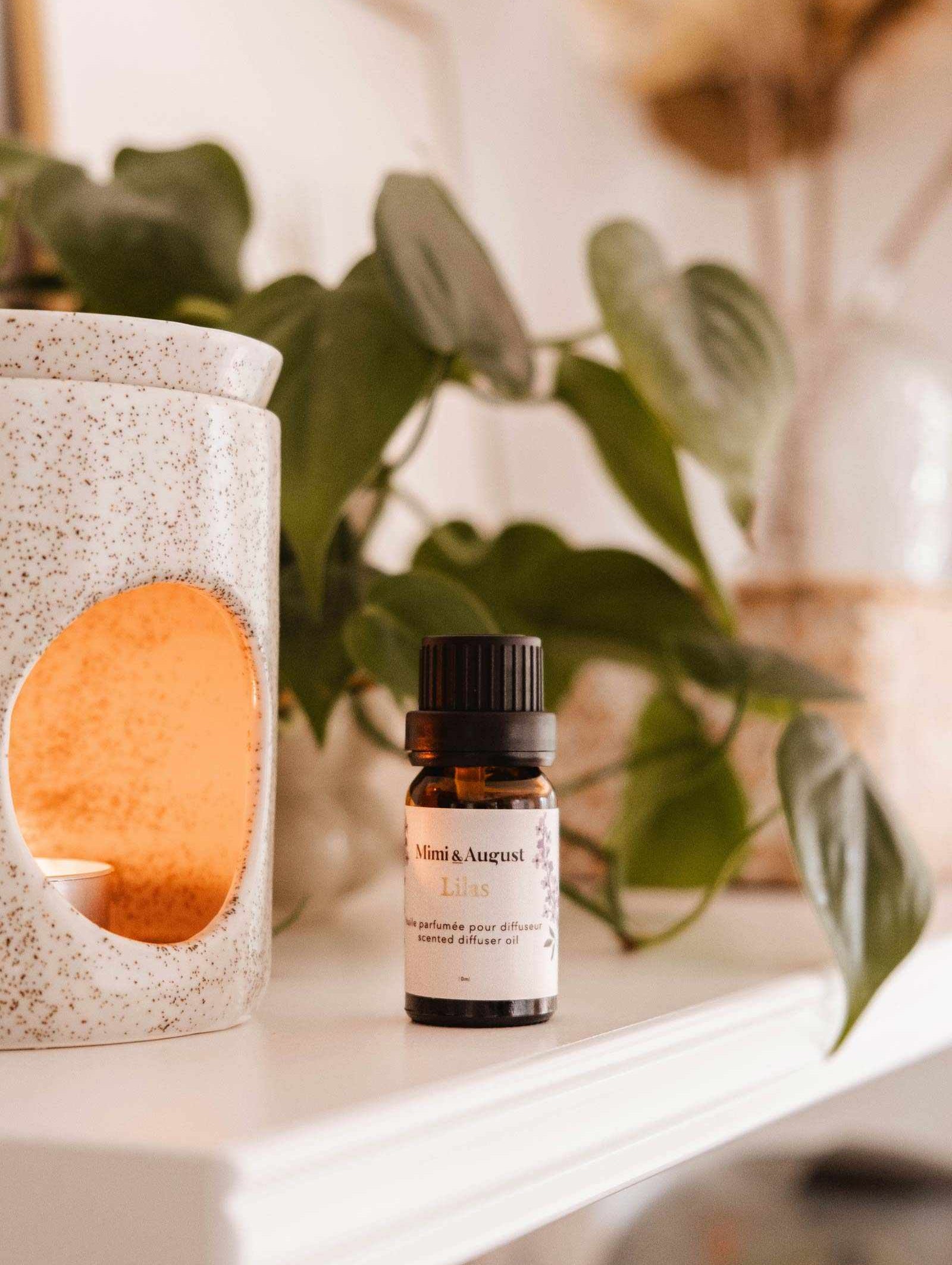A small bottle labeled "Mimi & August Scented Oil - Lilac" is placed on a shelf beside a white ceramic oil diffuser with a lit candle inside, releasing its calming and soothing lilac scent. In the background, green leaves and neutral decor are visible.
