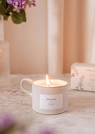 The Lilac - Reusable Candle by Mimi & August, in a white ceramic cup, sits on marble with a box and purple flowers—an inviting lilac scent and stylish touch for your home decor.