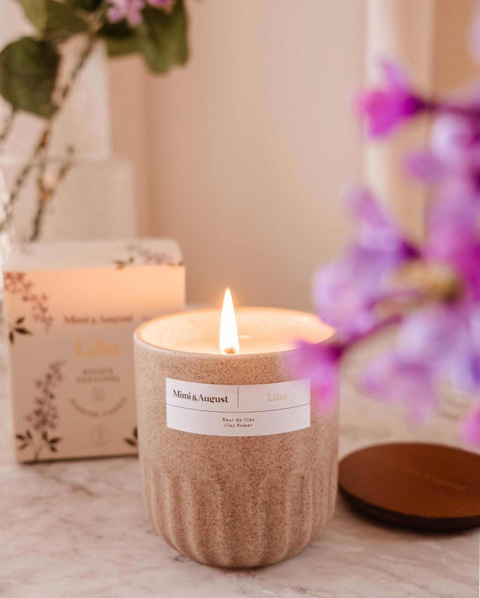 The Lilac - Reusable Candle by Mimi & August in a beige ceramic jar sits on marble, bringing aromatherapy and elegance to your home, with its box and purple flowers as a stylish backdrop.