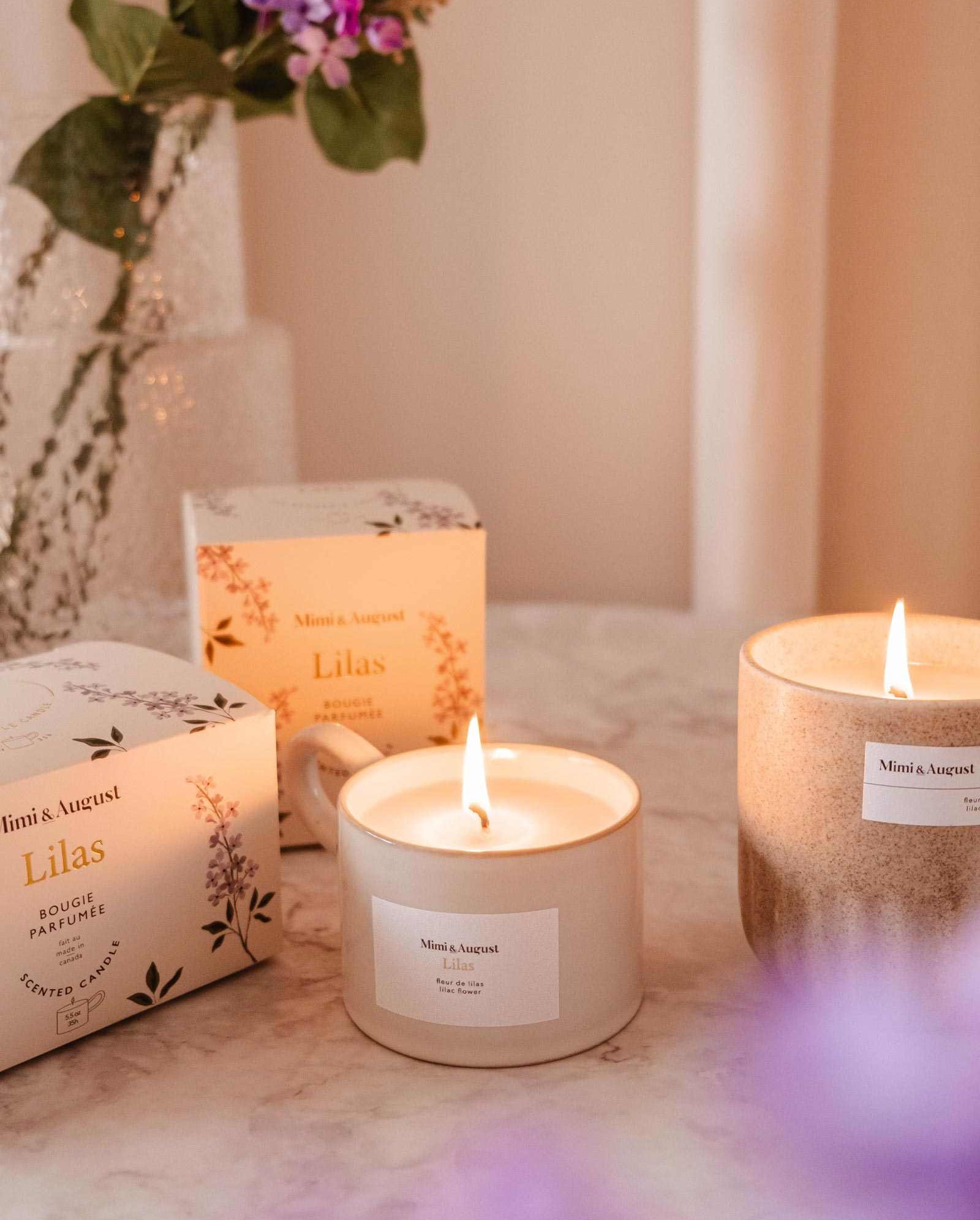 Two lit Mimi & August Lilac - Reusable Candles labeled "Lilas" sit on marble, with a vase of purple flowers in the background—ideal for adding soothing aroma and elegant home decor to your space.