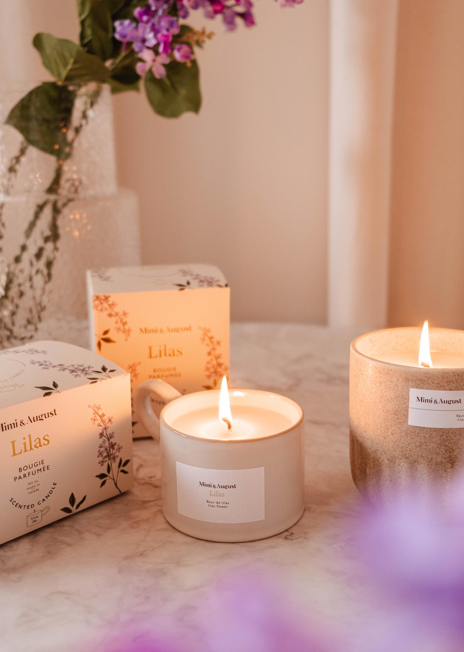 Two lit Mimi & August Lilac - Reusable Candles labeled "Lilas" sit on marble, with a vase of purple flowers in the background—ideal for adding soothing aroma and elegant home decor to your space.