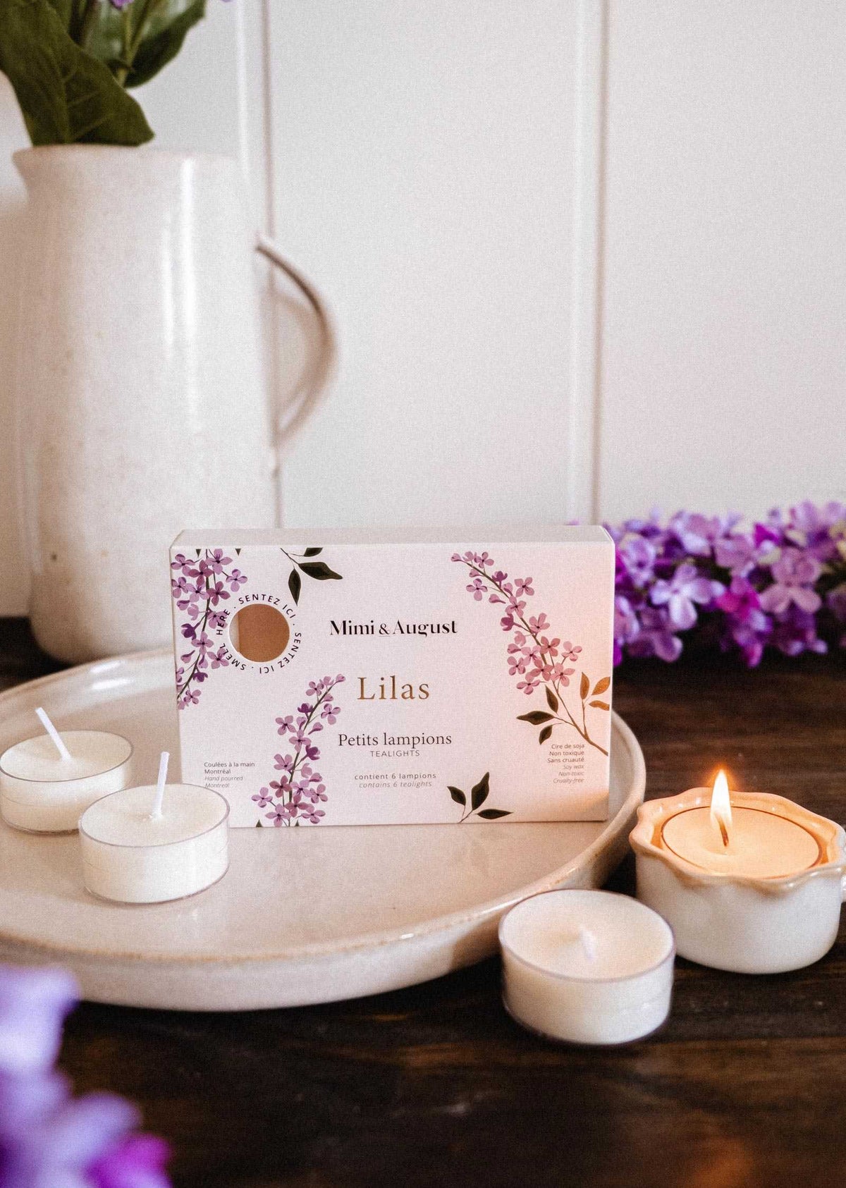 A Mimi & August Lilac Tealight Candle Set rests on a ceramic plate with four soy tealights—one lit—surrounded by lilac flowers and a white pitcher in the background.