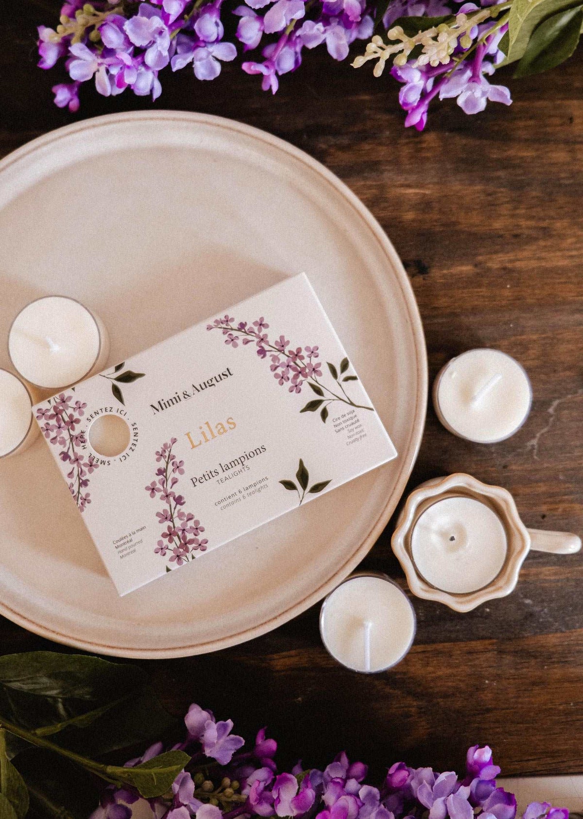 The Mimi & August Lilac Tealight Candle Set features a floral lilac box on a plate, surrounded by natural soy wax tealights, a ceramic holder, and purple flowers on a wooden surface.