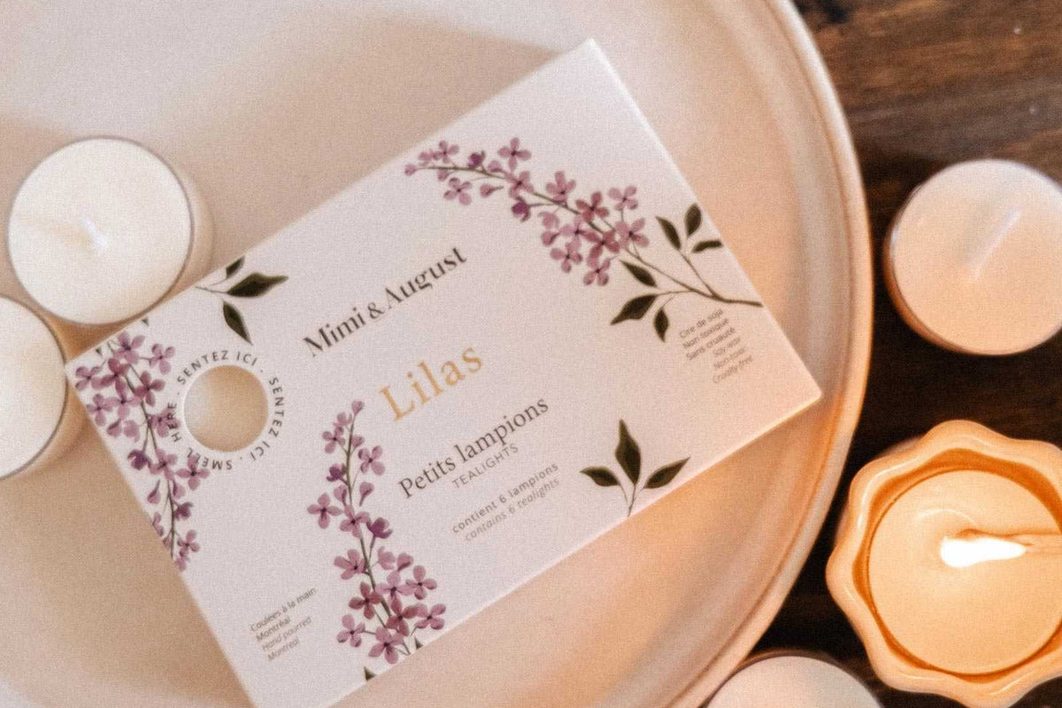 The Lilac Tealight Candle Set by Mimi & August, in a floral-designed box, is displayed on a plate with soy tealights and purple flowers arranged on a wooden surface.