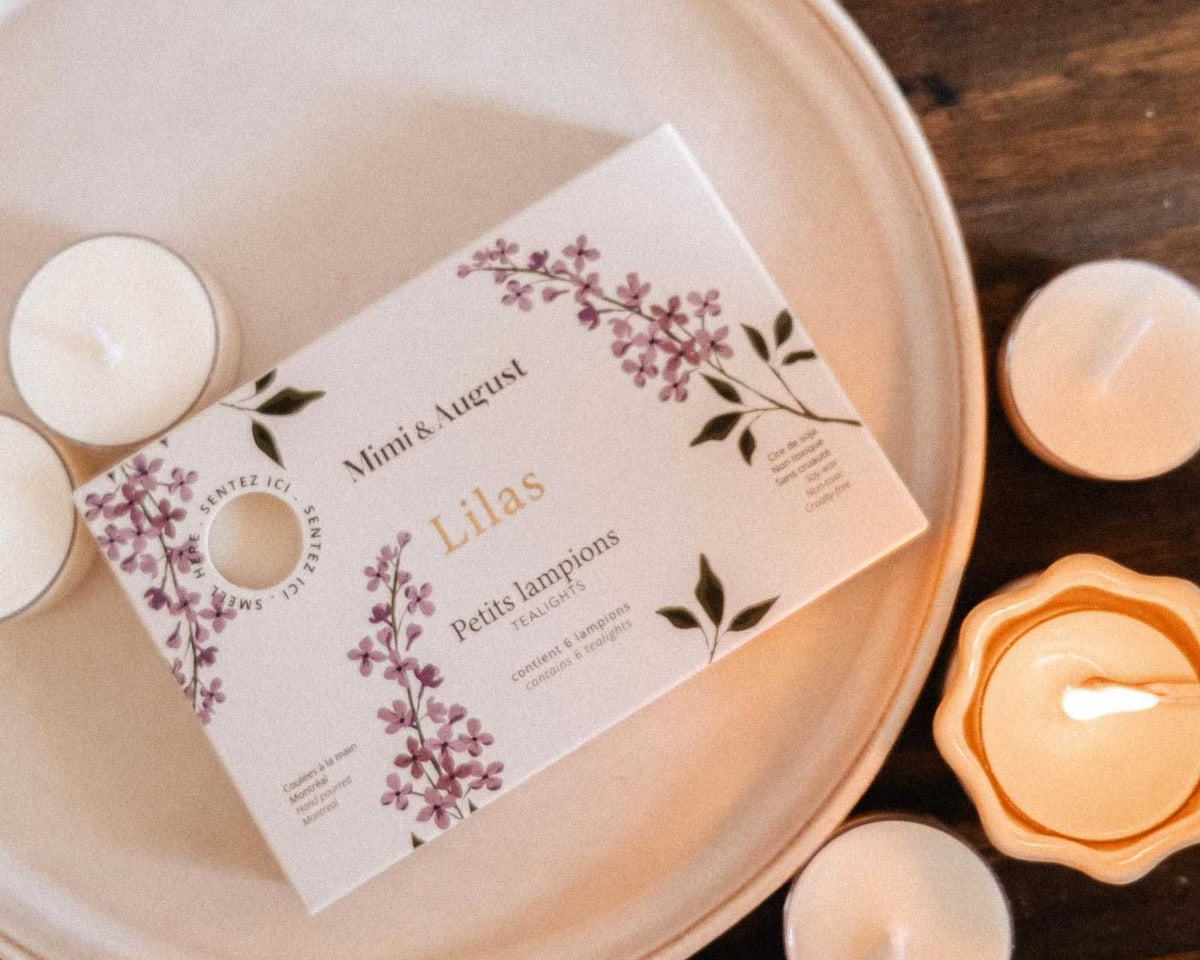 Le Coffret de lampions parfumés Lilas de Mimi & August, dans une boîte au design floral, est présenté sur une assiette avec des lampions en cire de soya et des fleurs violettes disposées sur une surface en bois.