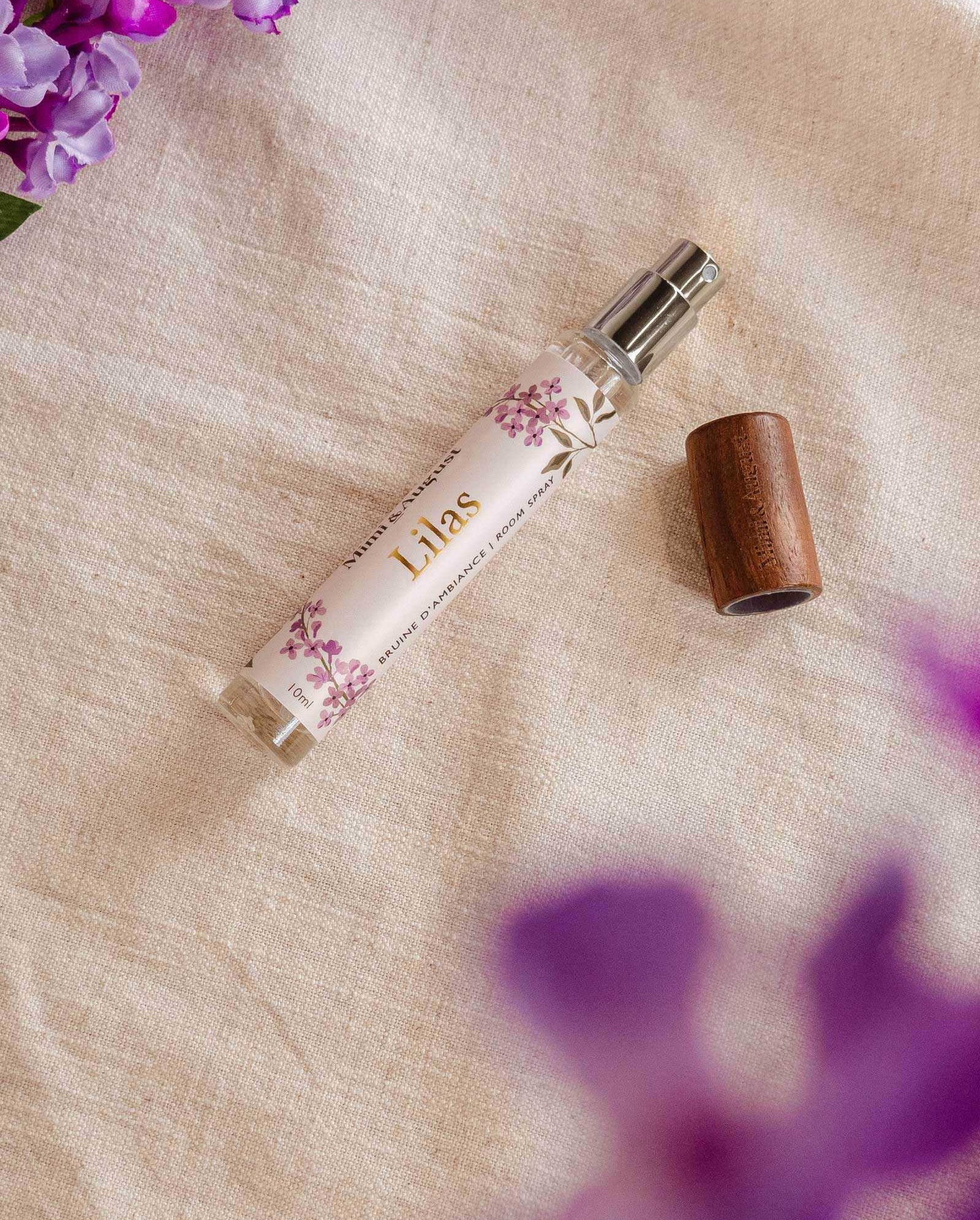 A spray bottle of Mimi & August Room spray - Lilas lies on a fabric surface with a wooden cap next to it. There are purple flowers near the top left and bottom right corners of the image, exuding a touch of lilac fragrance.