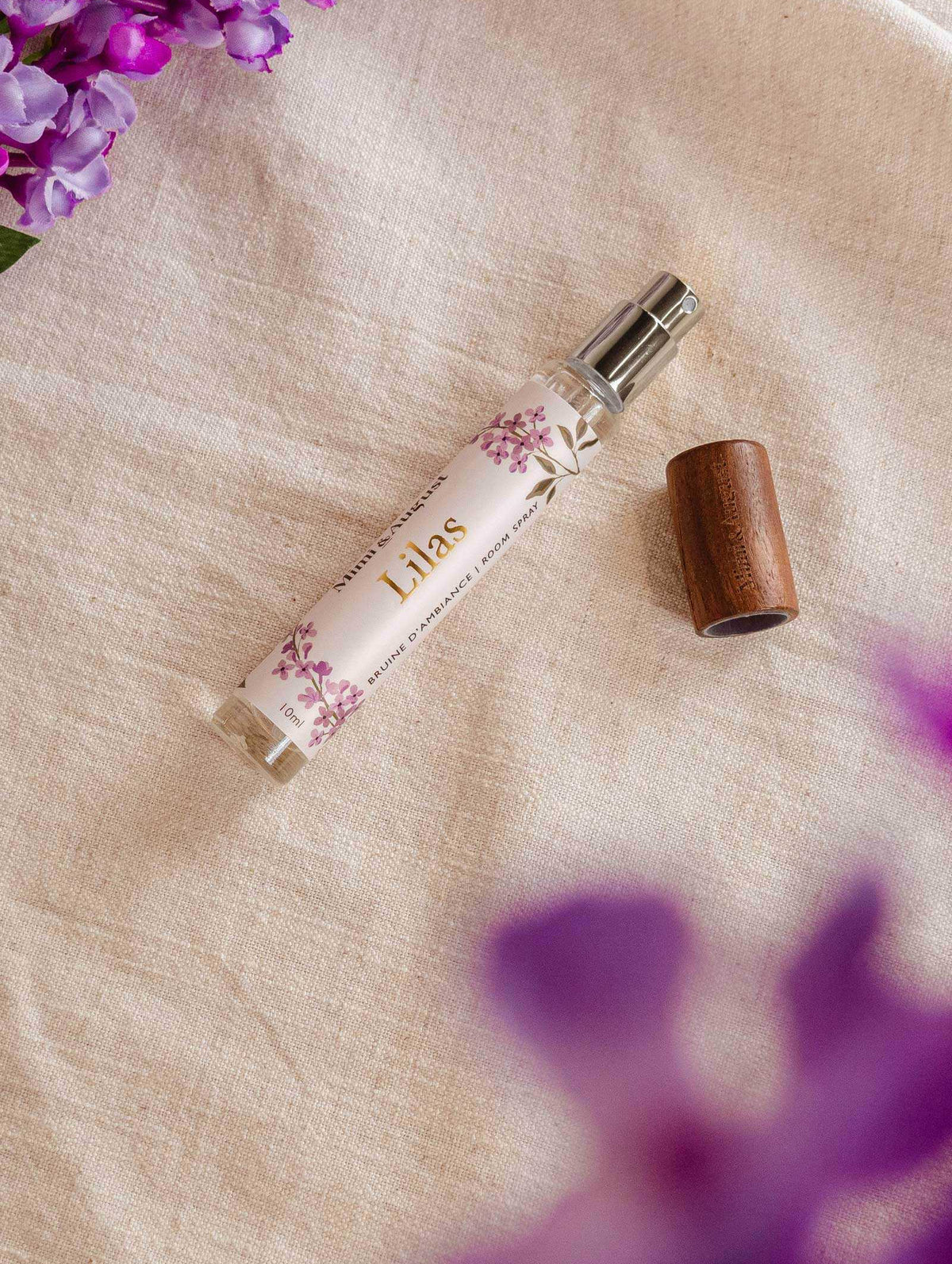 A spray bottle of Mimi & August Room spray - Lilas lies on a fabric surface with a wooden cap next to it. There are purple flowers near the top left and bottom right corners of the image, exuding a touch of lilac fragrance.