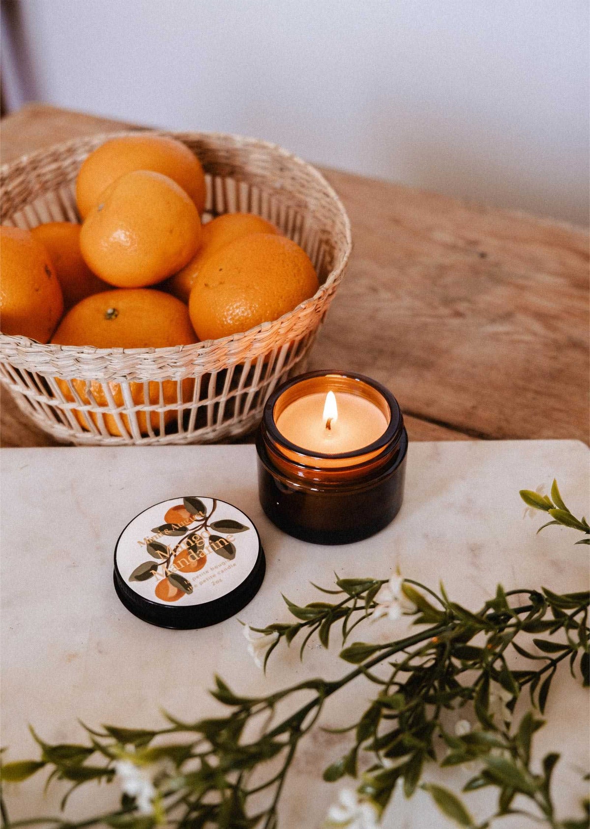 A lit Mimi & August Mango Mandarine – 2 oz Soy Candle in a brown glass jar sits on a table next to a basket of oranges, its lid, and green foliage.