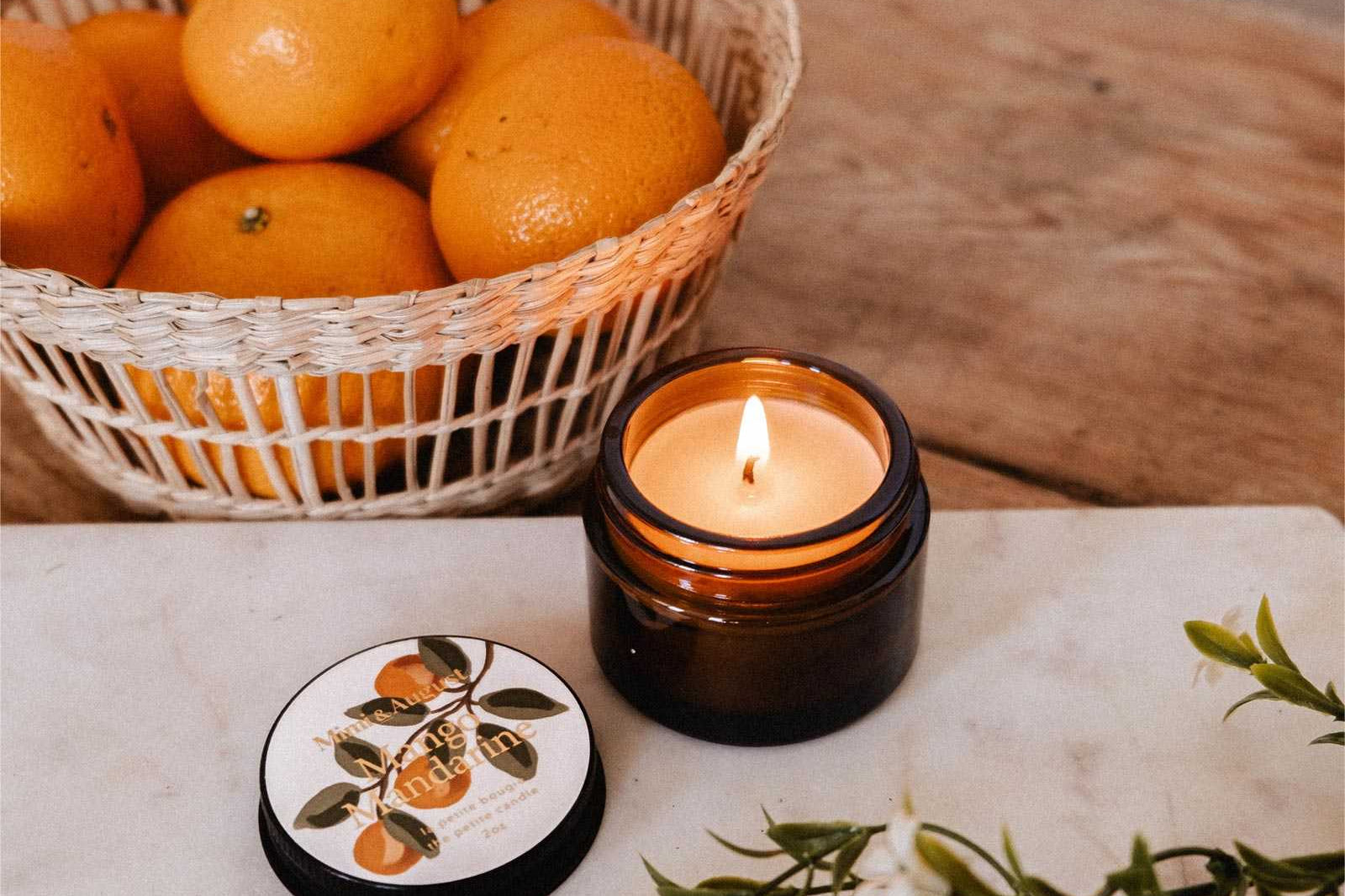 A lit Mimi & August Mango Mandarine – 2 oz Soy Candle in a brown glass jar sits on a table next to a basket of oranges, its lid, and green foliage.