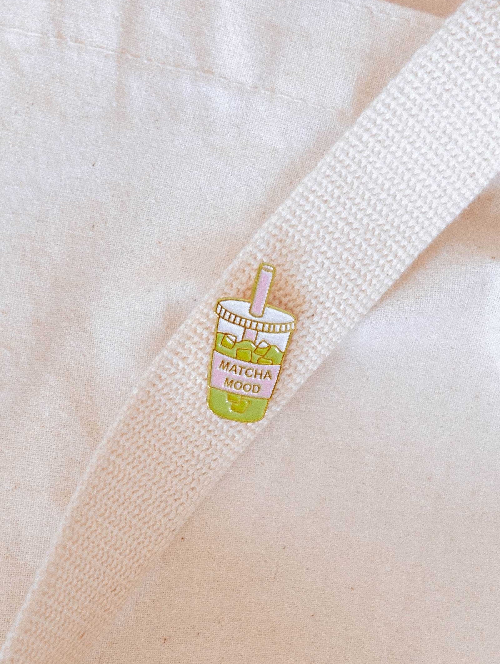 The Matcha Mood Enamel Pin by Mimi & August is a cute cup of matcha bubble tea with "MATCHA MOOD" text, attached to a white cloth bag—an ideal gift for any matcha lover.