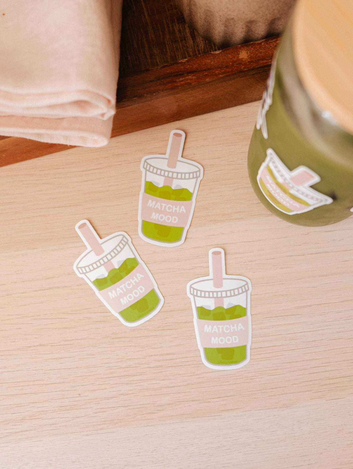 Three Mimi & August "Matcha Mood - Vinyl Sticker" durable iced matcha drink-shaped stickers rest on a light wooden surface beside a jar with the same sticker and a folded beige napkin.