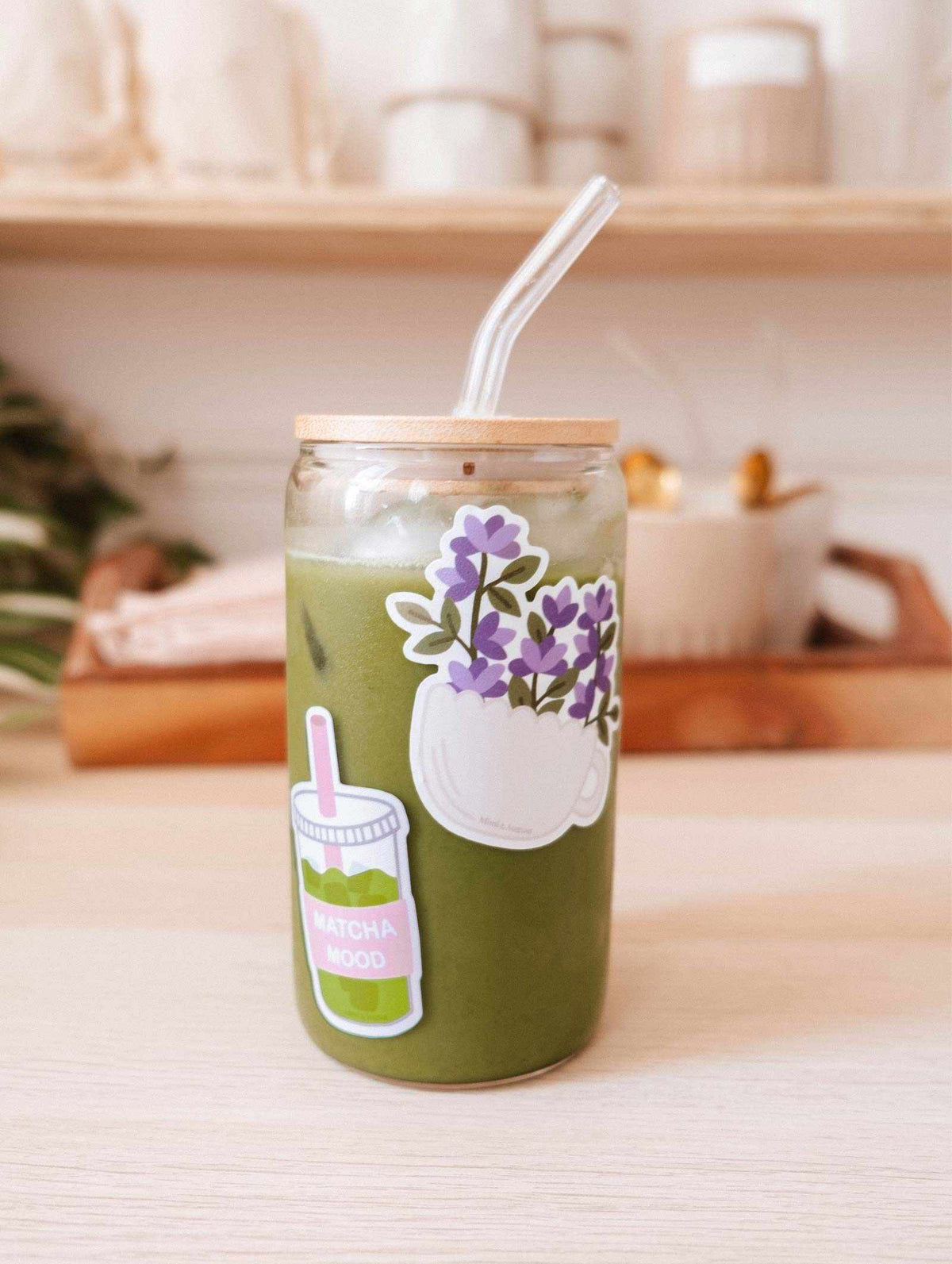 A glass jar of iced matcha latte with a bamboo lid, clear straw, and the Matcha Mood - Vinyl Sticker by Mimi & August sits on a light wooden table, with blurred kitchen items visible in the background.