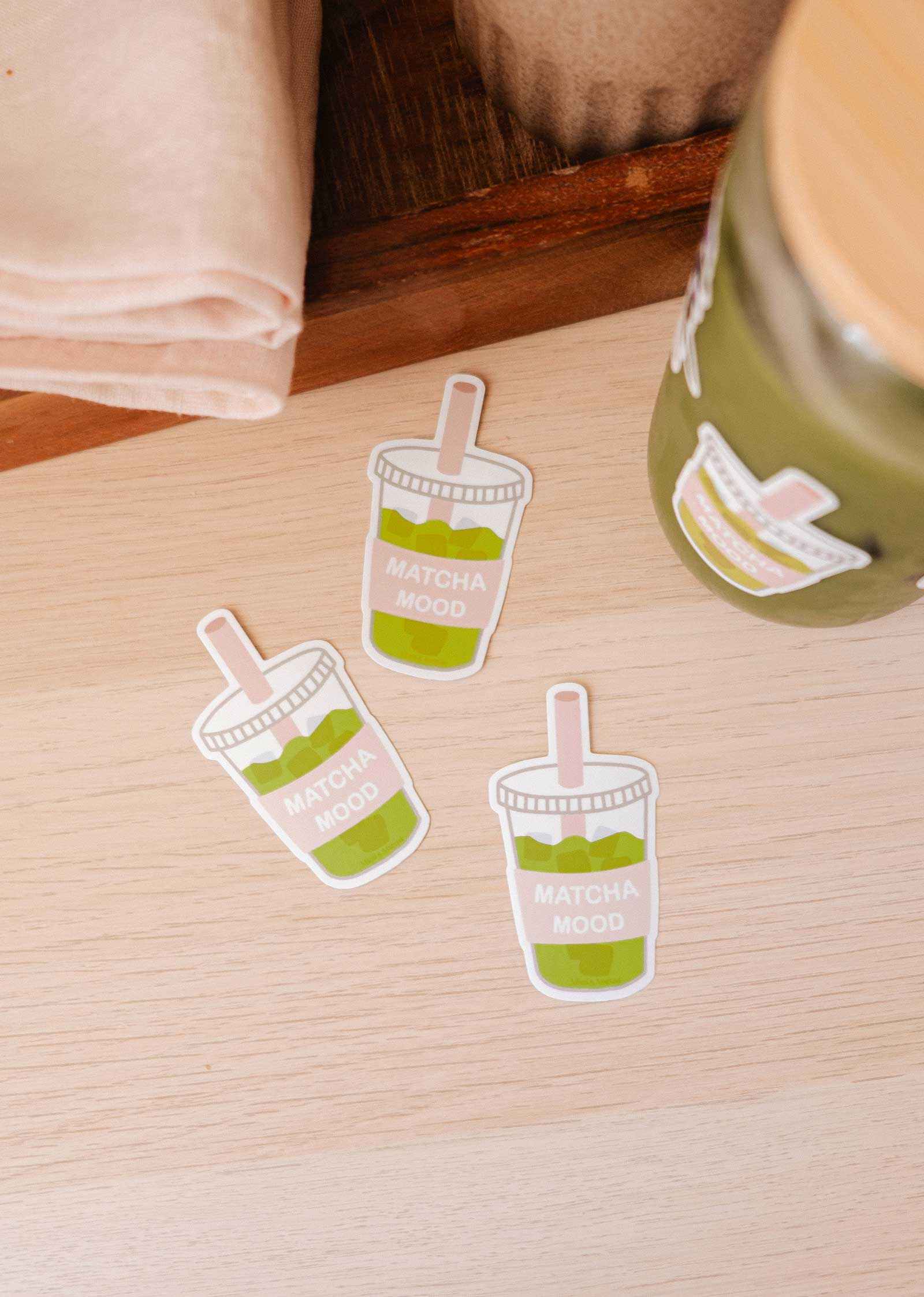 Three Mimi & August "Matcha Mood - Vinyl Sticker" durable iced matcha drink-shaped stickers rest on a light wooden surface beside a jar with the same sticker and a folded beige napkin.