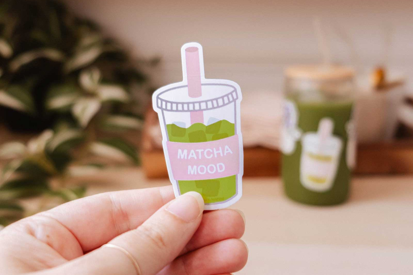 A hand holds the "Matcha Mood - Vinyl Sticker" by Mimi & August, featuring a cartoon iced matcha and the label "MATCHA MOOD," with a blurred green cup and shelves in the background.