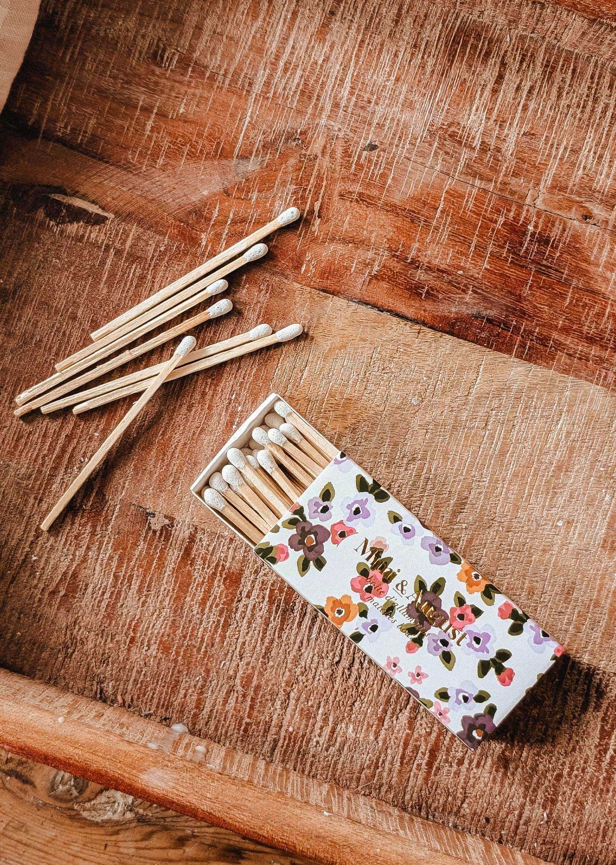 The Mimi & August Mimi Floral Matchbox - 30 Sticks, filled with eco-friendly matches, sits on a wooden surface with several matches scattered nearby.