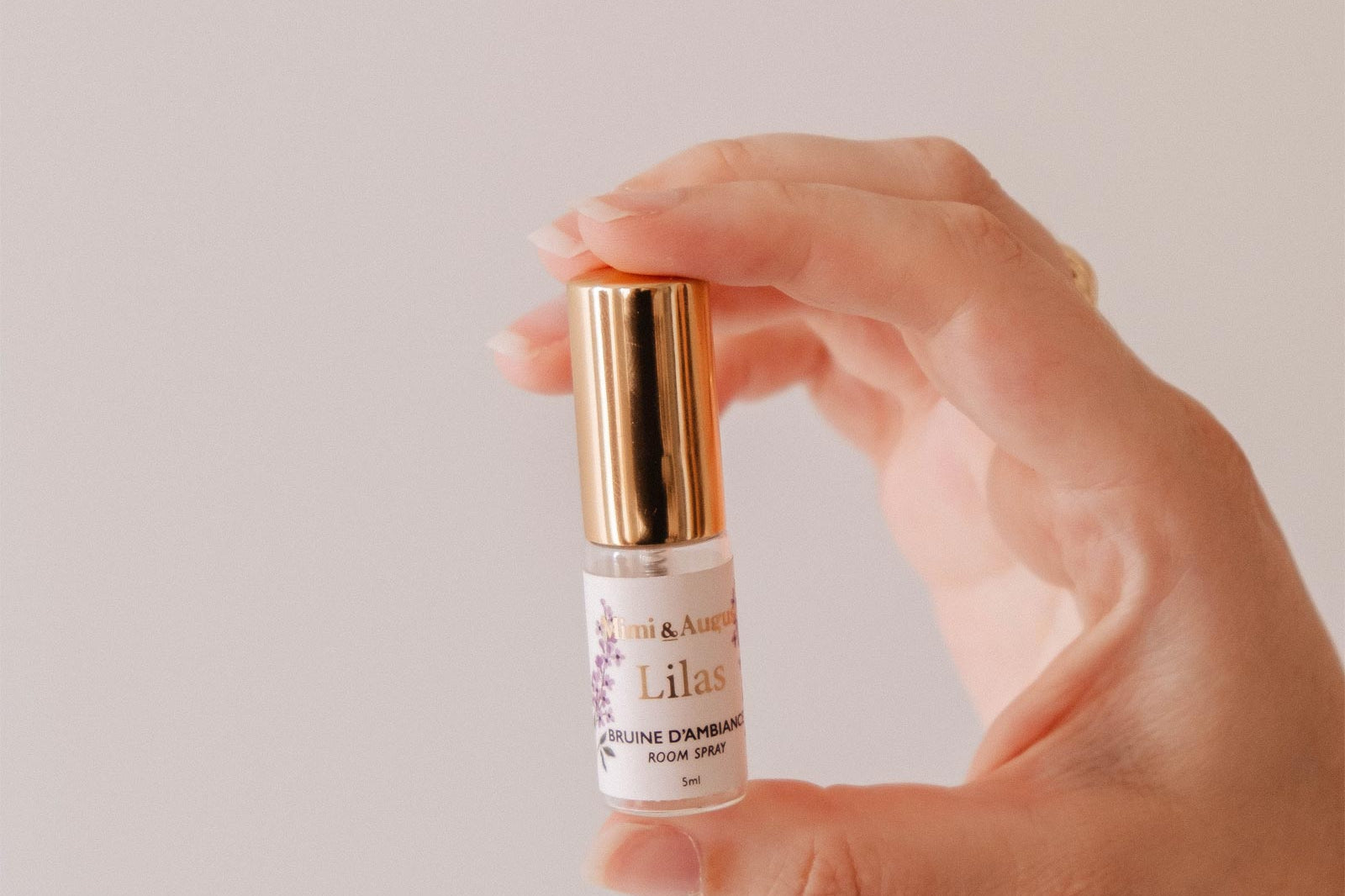 A hand holds a Mini Room spray - Lilas by Mimi & August, featuring a gold cap and label, against a plain light background—the perfect way to enjoy a delightful lilac fragrance.