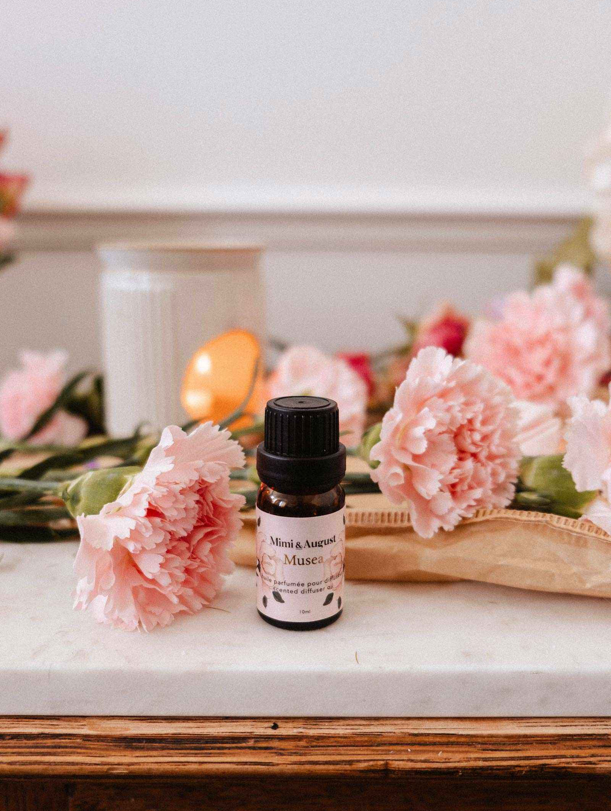 Une petite bouteille d'huile parfumée Musea de Mimi & August repose sur une surface en marbre, avec des œillets roses, une bougie blanche et un livre à proximité.