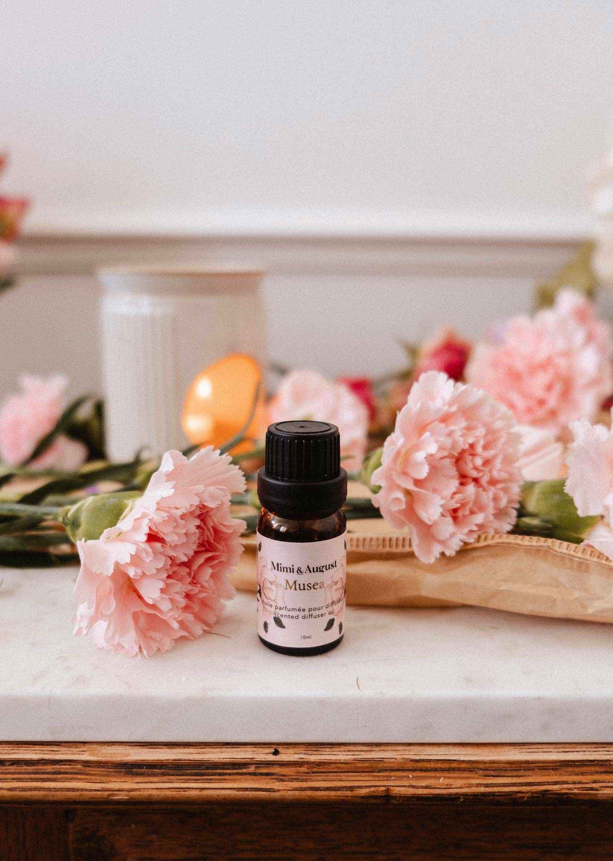 Une petite bouteille d'huile parfumée Musea de Mimi & August repose sur une surface en marbre, avec des œillets roses, une bougie blanche et un livre à proximité.