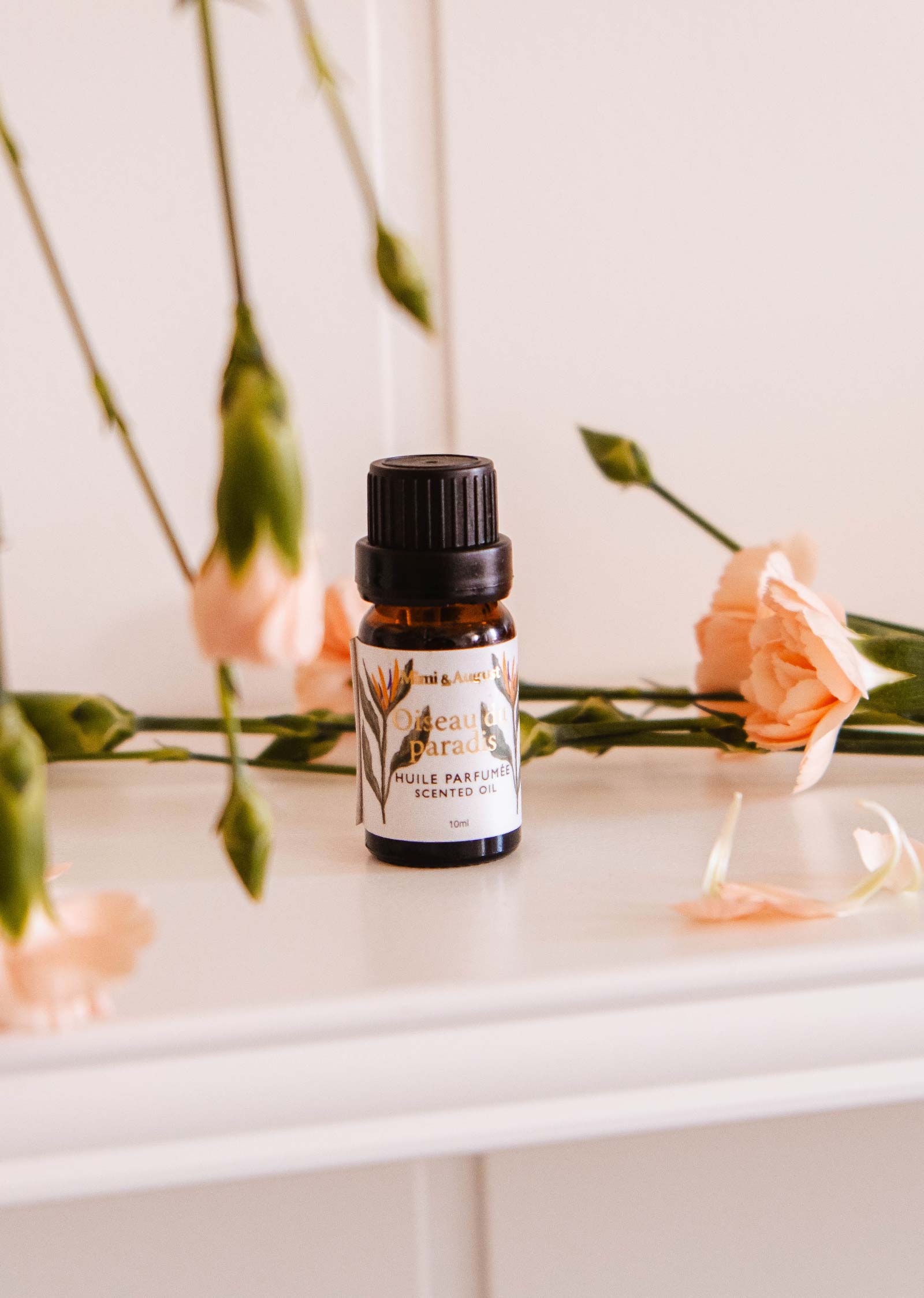 A small amber bottle of Mimi & August's Scented Oil - Oiseau du paradis sits on a white surface, surrounded by pink carnations and green stems—ideal for use as a diffuser oil.