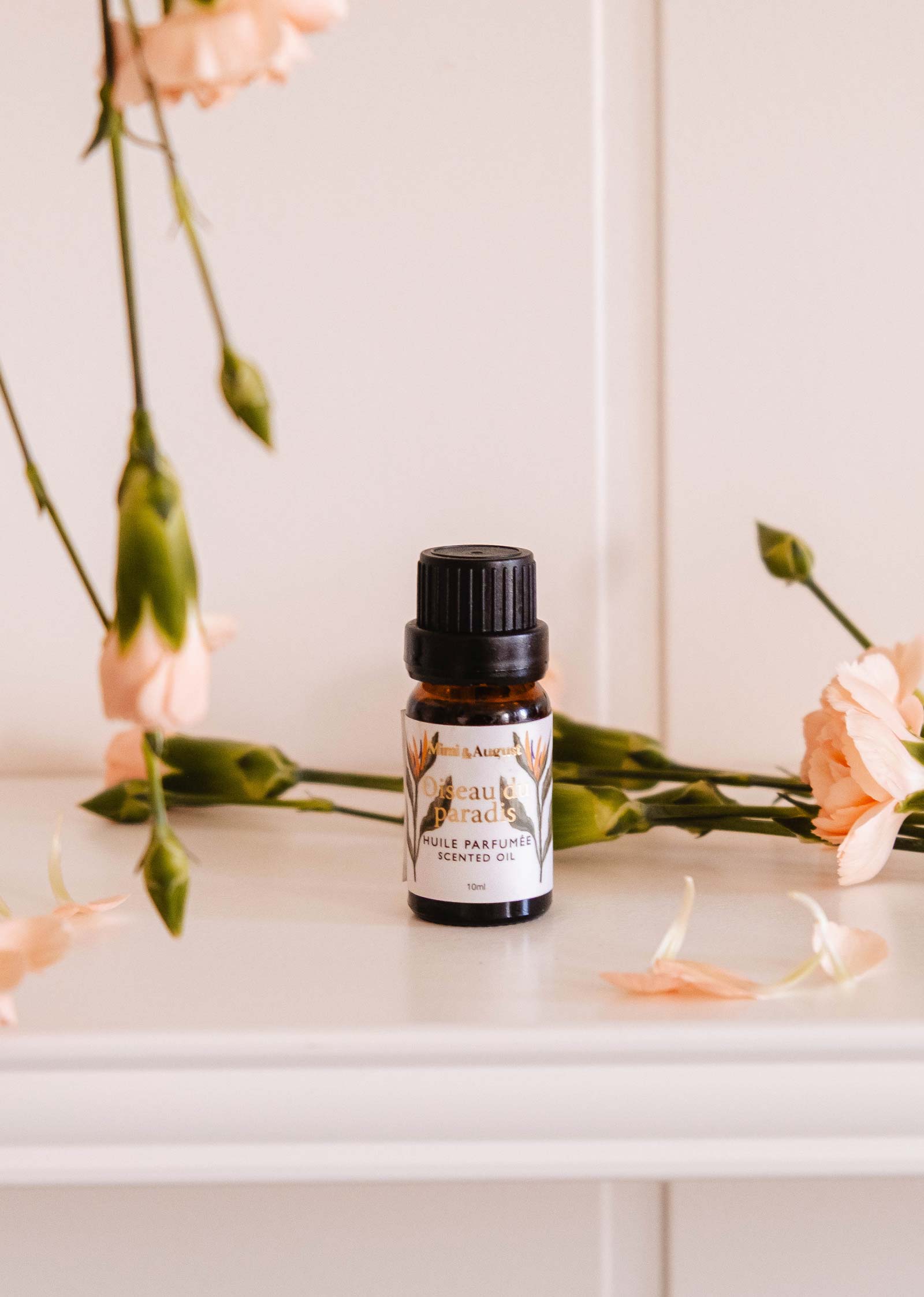 A small brown bottle of Mimi & August's Scented Oil - Oiseau du paradis, featuring a floral label, sits on a white surface surrounded by light pink flowers and green stems.