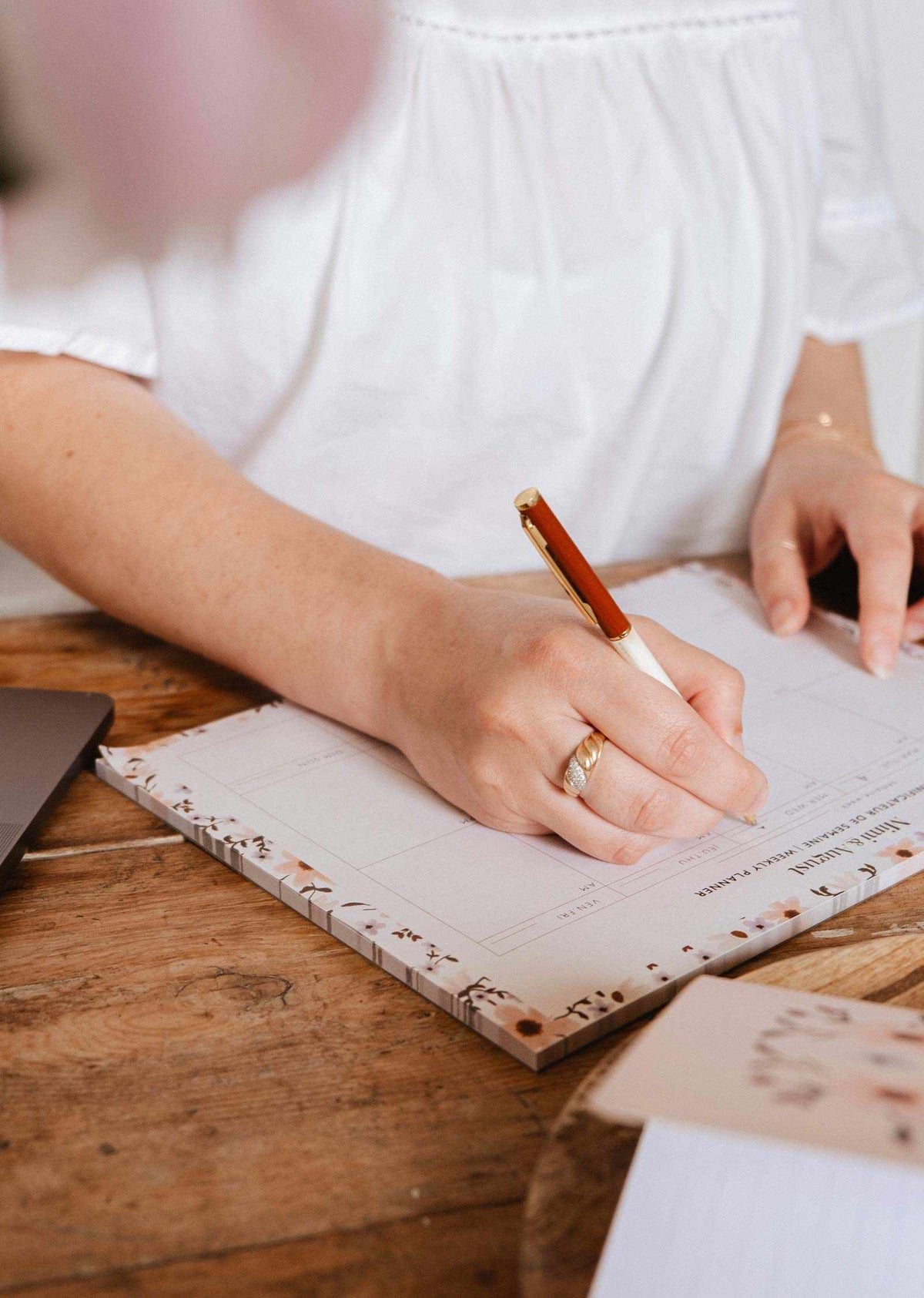 A person writes with a pen on the Mimi & August Petals - Weekly Planner at a wooden desk next to a laptop, staying organized and productive.