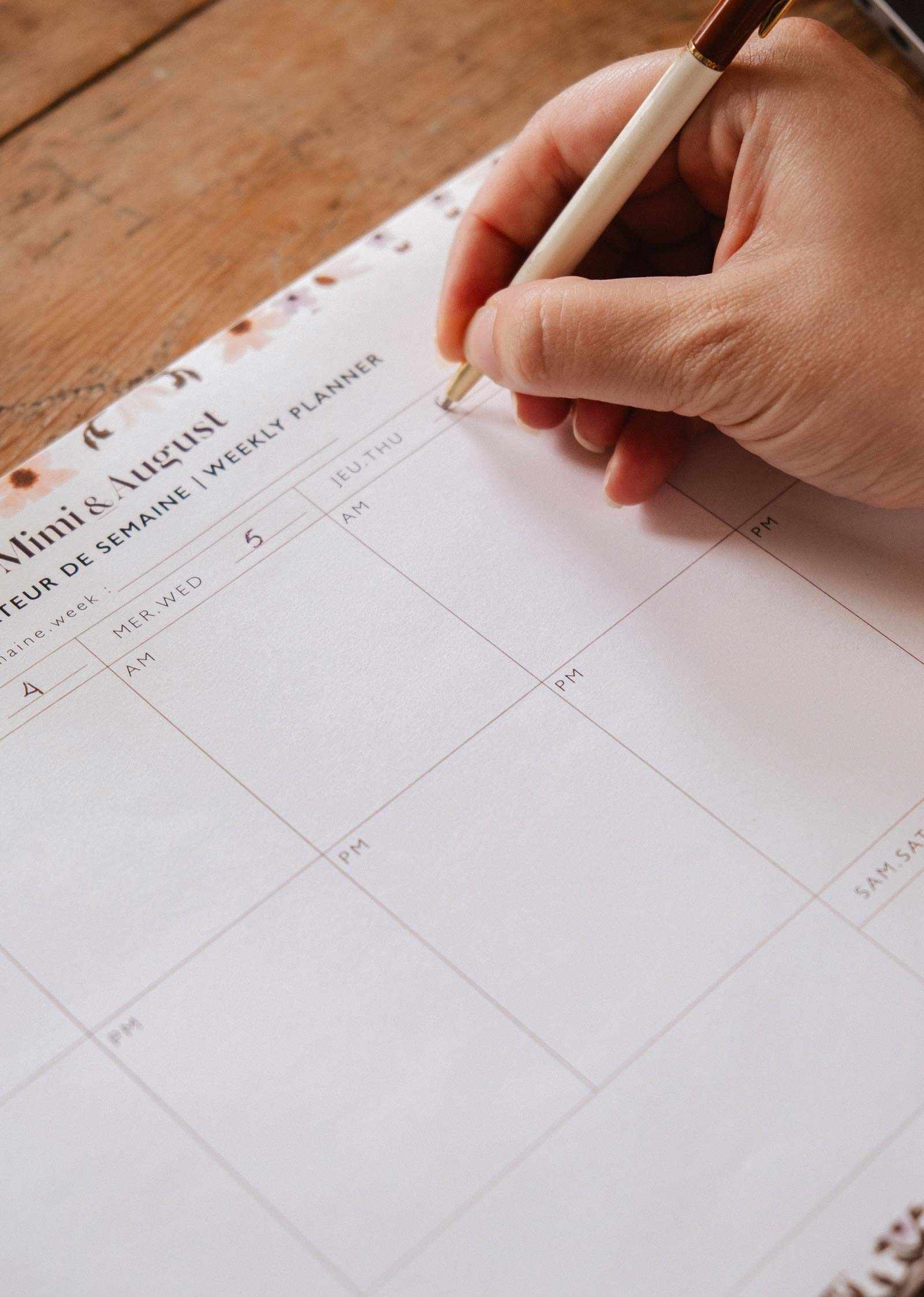 A hand holding a pen writes on a blank Petals - Weekly Planner by Mimi & August, made of recycled paper and set on a wooden surface.