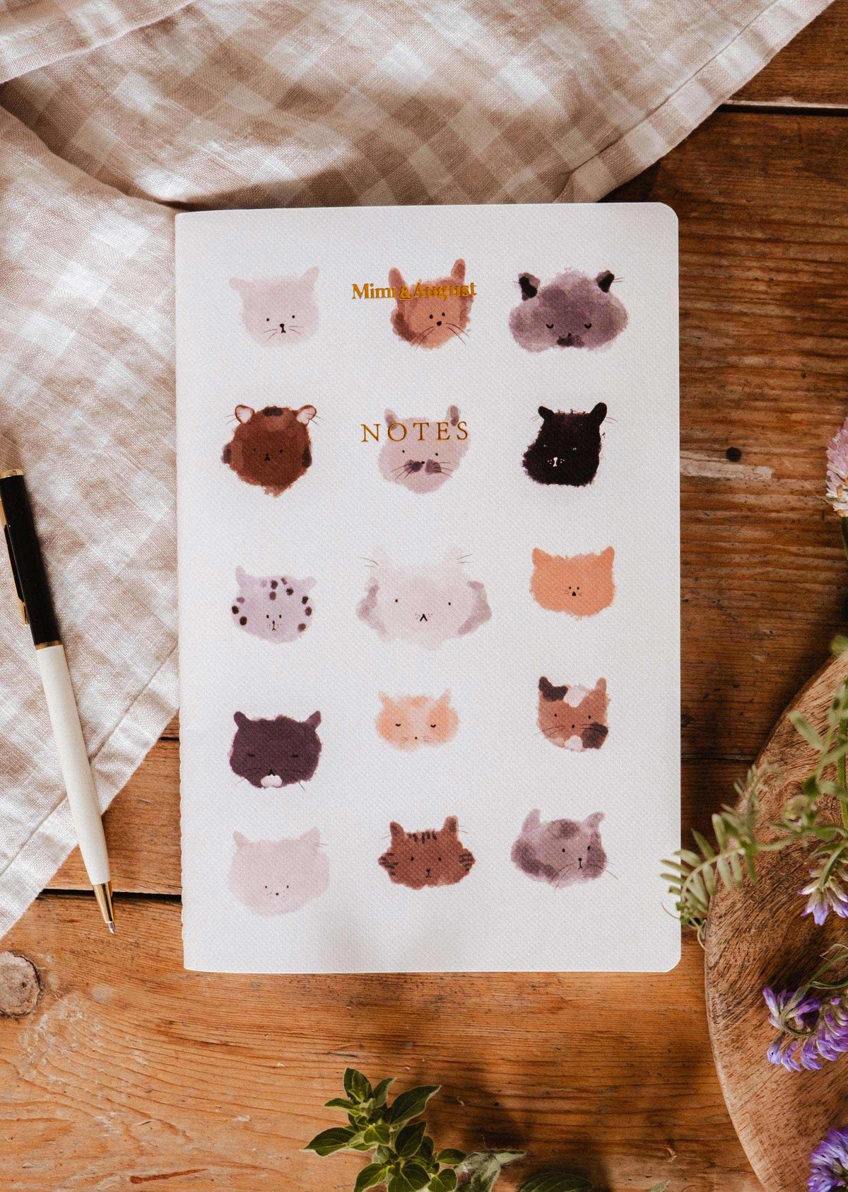 The Purrfect - Notebook by Mimi & August features illustrated cat faces on the cover and is shown on a wooden table next to a pen, checkered cloth, and flowers.