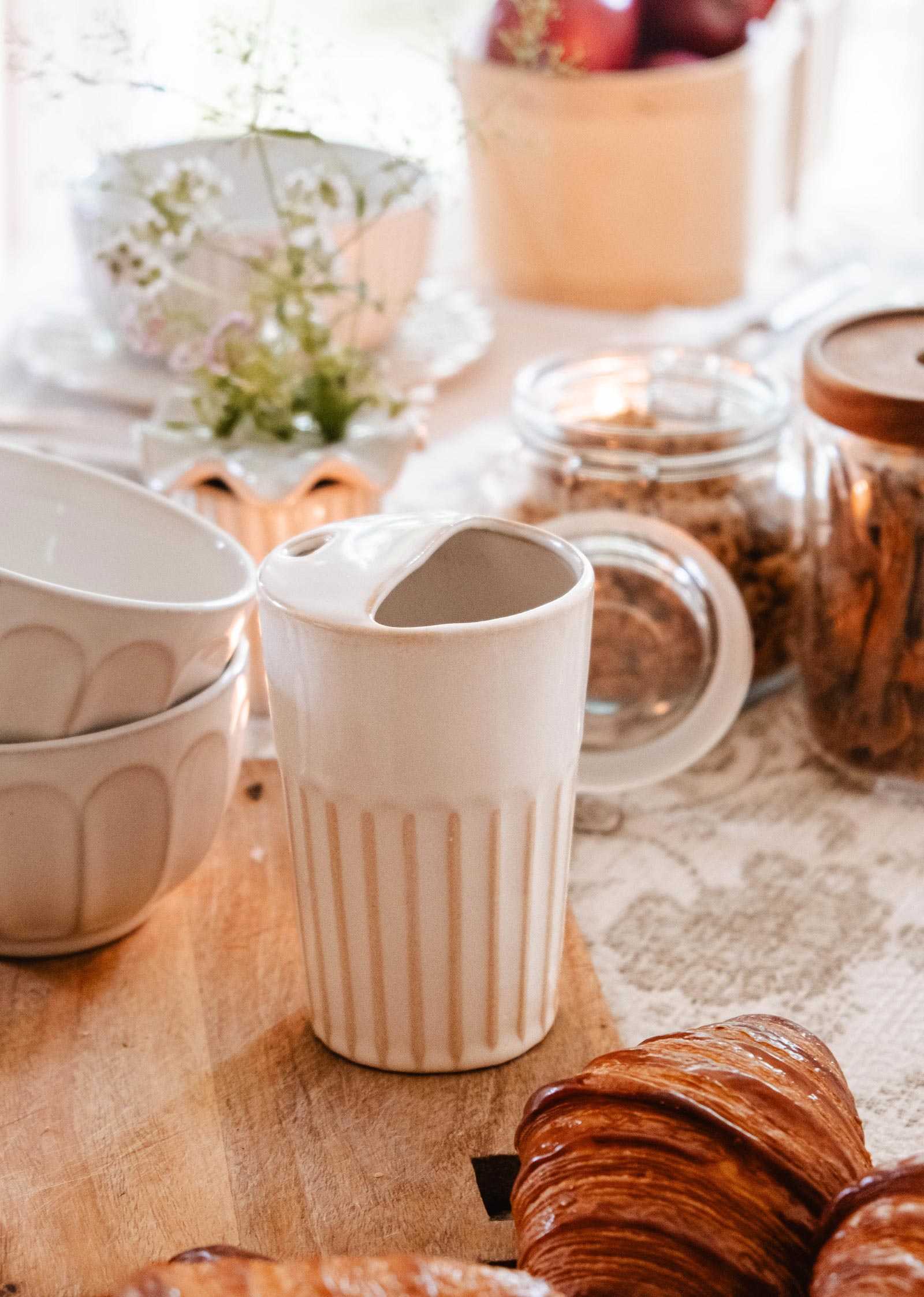 A white ceramic creamer sits on a wooden table surrounded by bowls, jars, a floral arrangement, croissants, and The Ribbed To-Go Cup by Mimi & August, which adds an artisanal touch to the cozy setting.